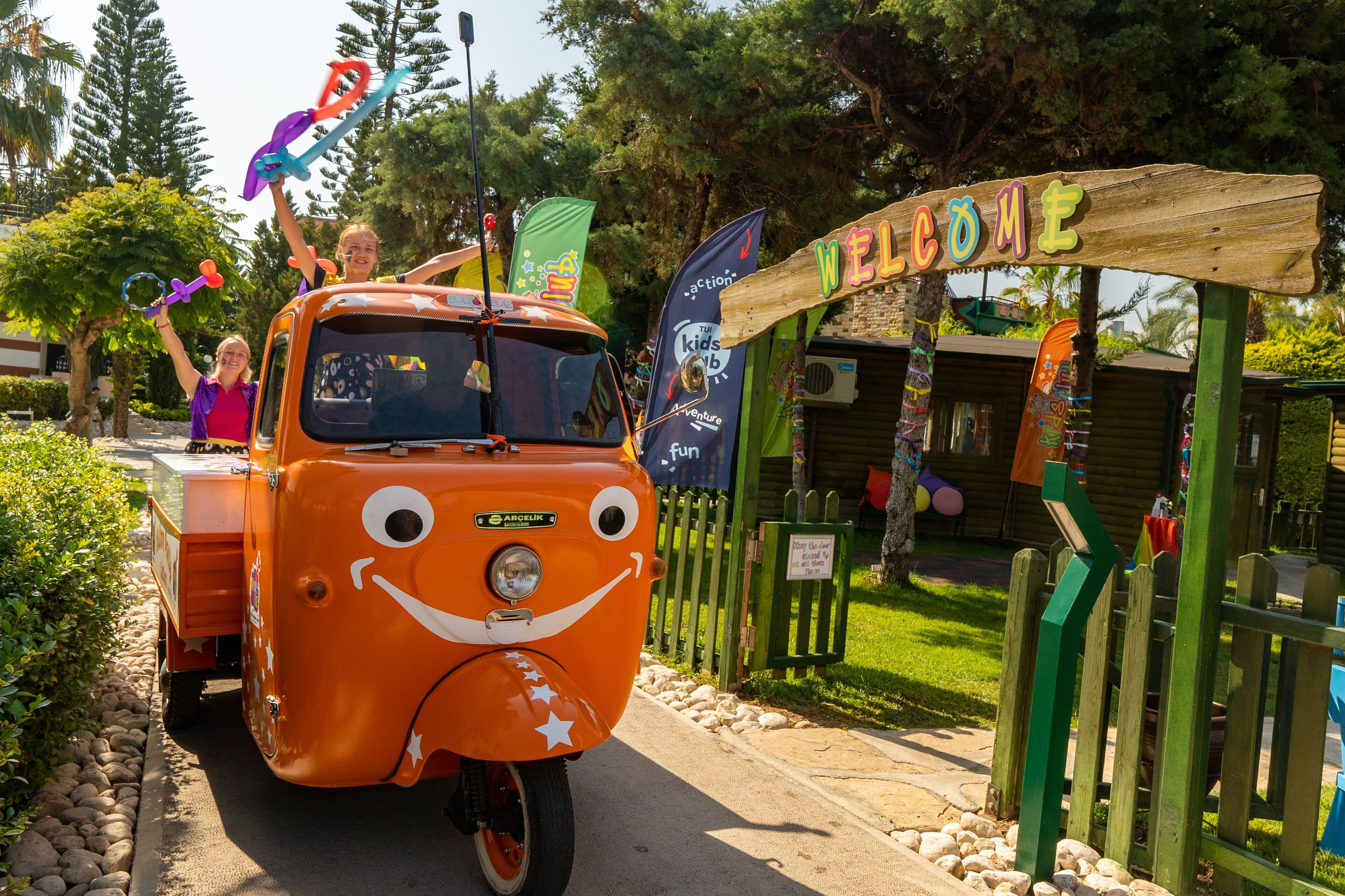 Ein orangefarbener Oldtimer mit einem freundlichen Gesicht und Sterndekorationen steht vor einem Holzschild mit der Aufschrift 'WELCOME'.