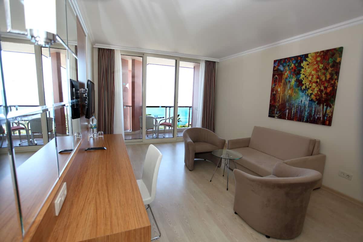 A modern hotel room with a wooden desk, a beige sofa, a glass coffee table, and a large window with a view of the sea.