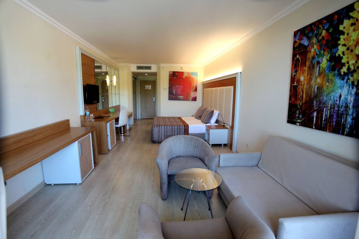 A modern hotel room with a living area featuring a sofa, armchairs, a glass coffee table, and a TV. There is a painting on the wall and a door leading to another room.