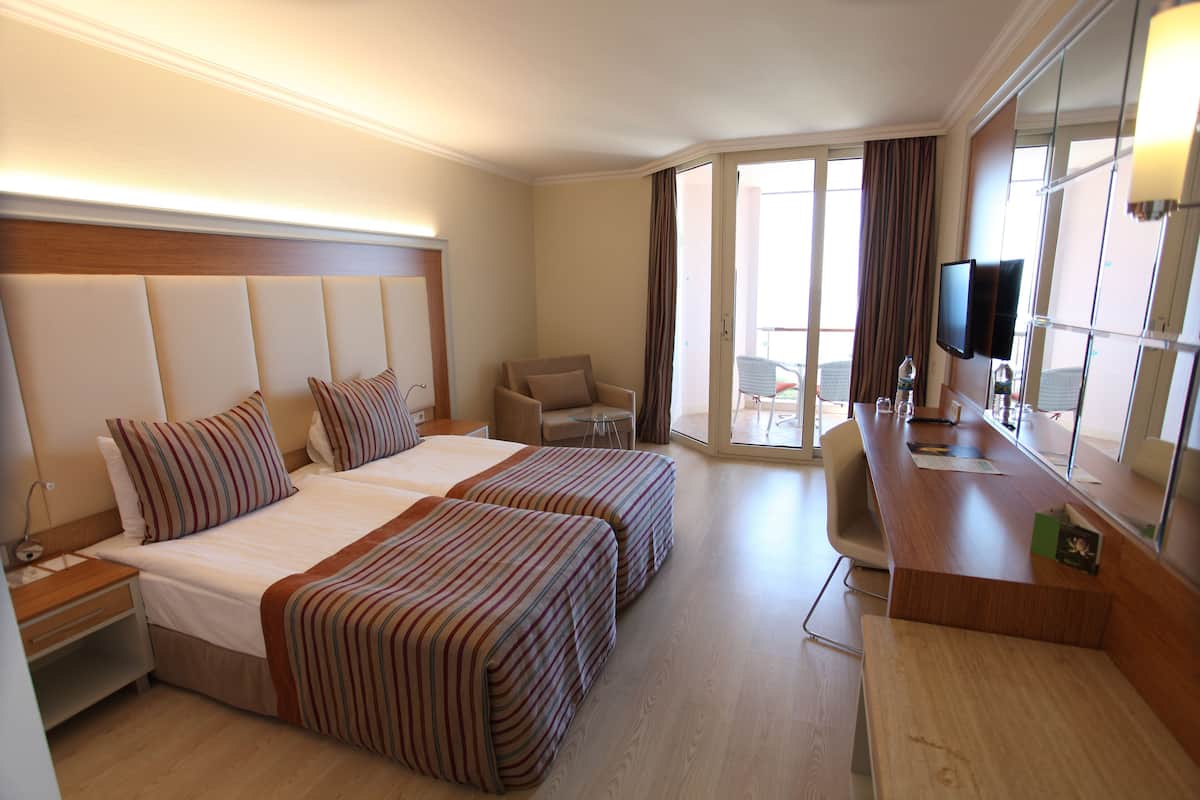 A hotel room with two beds, a desk, a chair, a TV, and a balcony with a table and chairs.