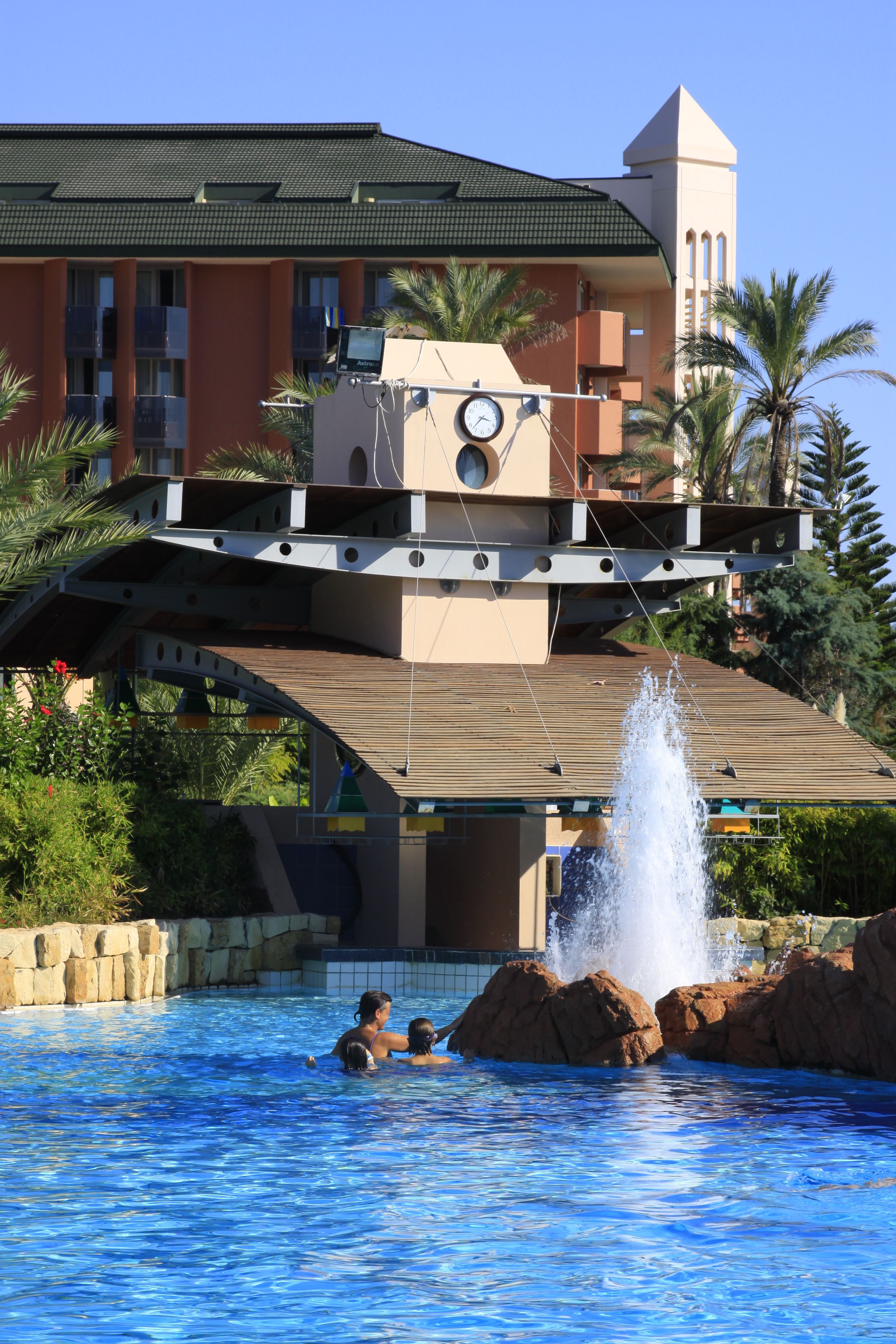 A swimming pool with a water fountain, palm trees, and a building with a tower in the background.