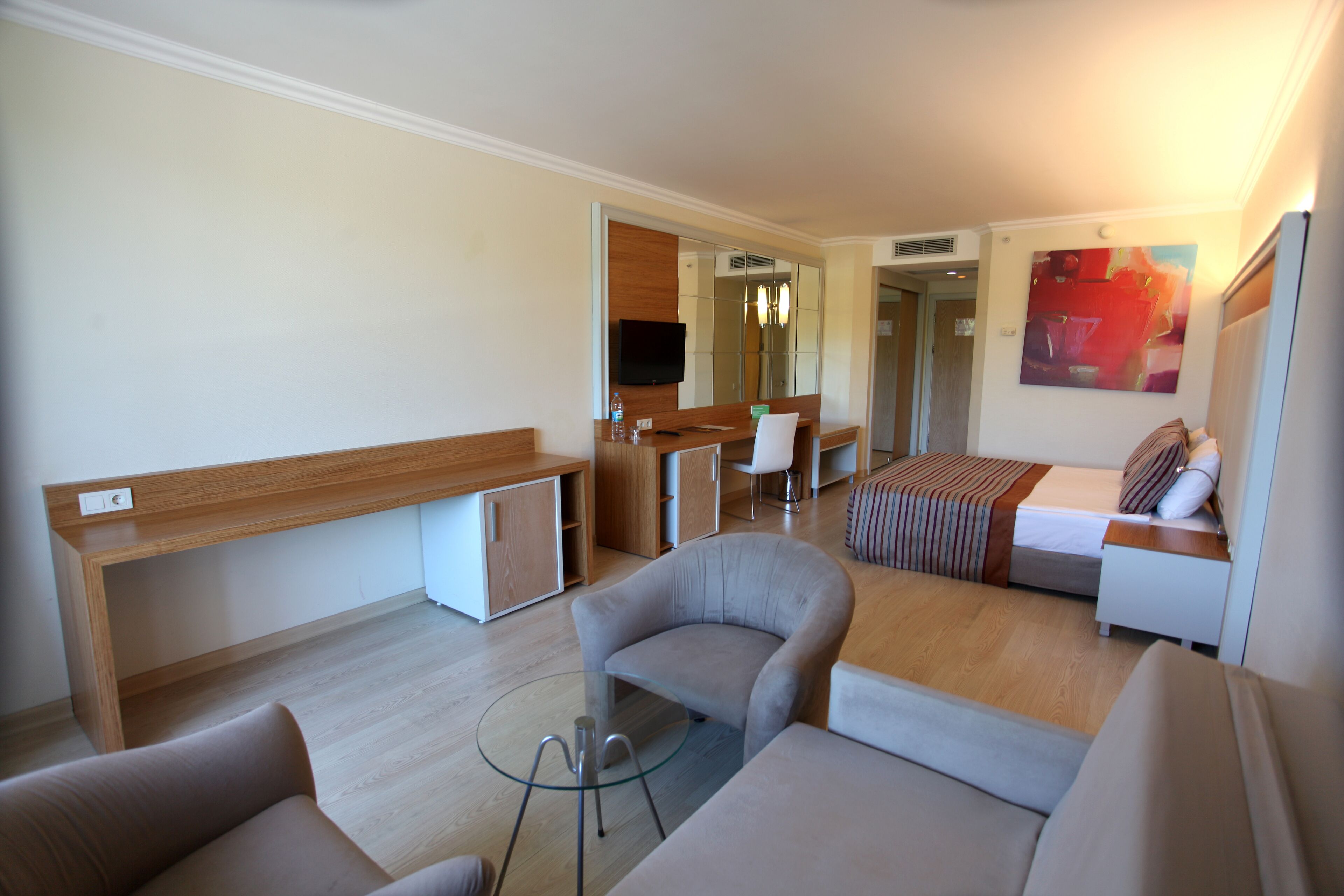A modern hotel room with a sofa, armchair, and a small table. There is a bed with a striped blanket and a TV mounted on the wall.