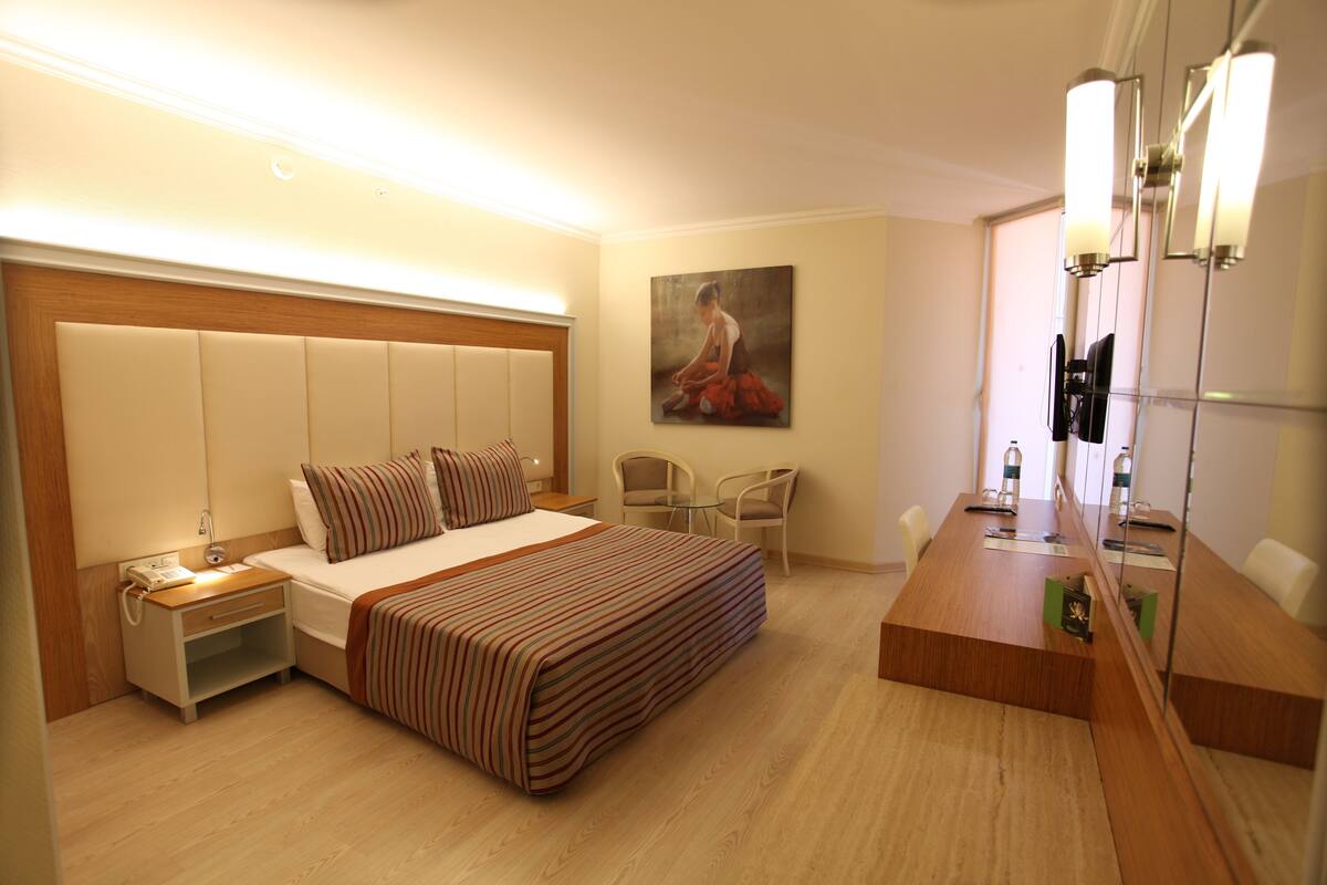 A hotel room with a large bed, bedside table, nightstand, and a mirror with lights.