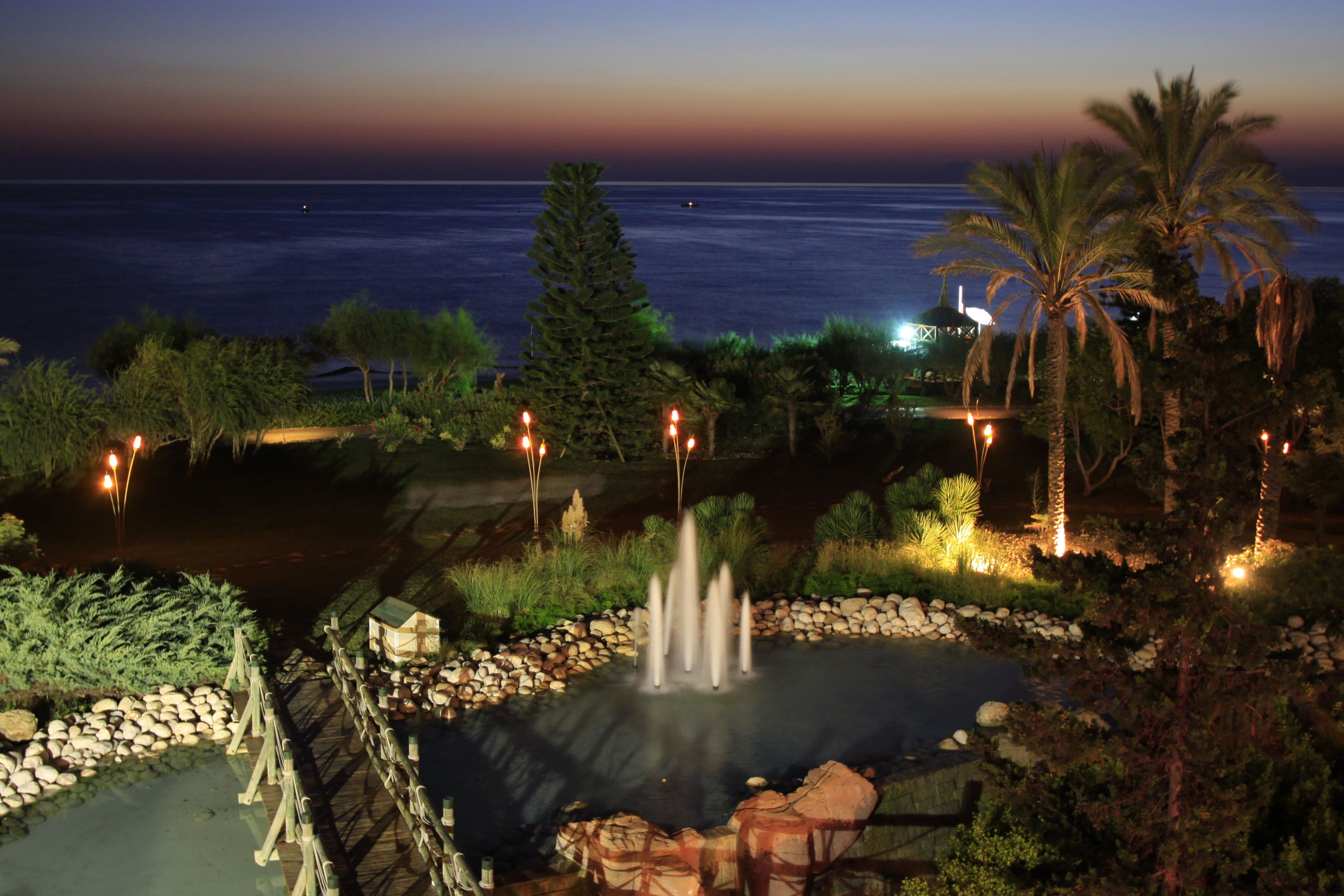 Vista nocturna de un jardín ajardinado con fuente de agua, palmeras y una vista distante del océano.