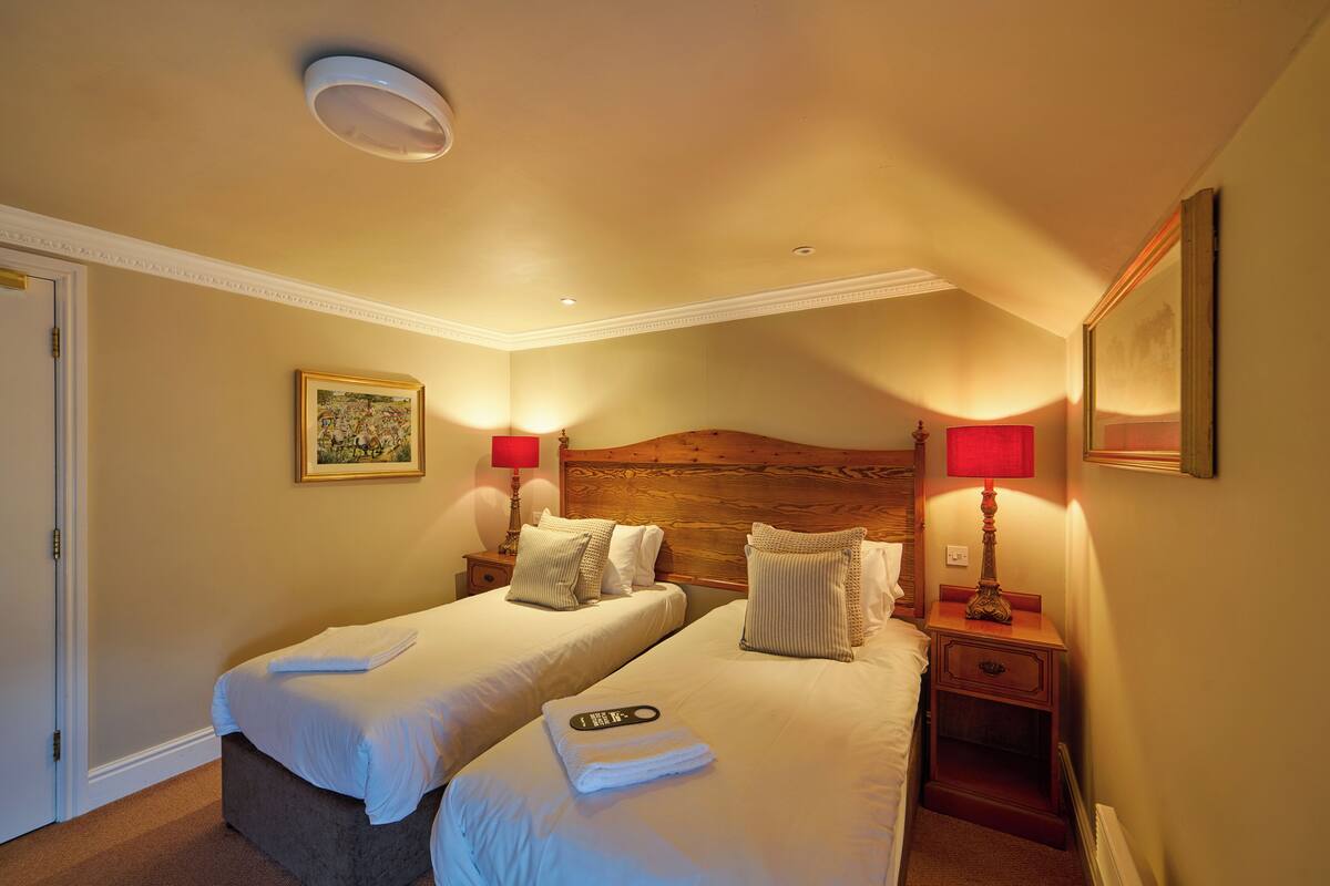 Une chambre d’hôtel avec deux lits, une tête de lit en bois, des lampes de chevet, un tableau encadré au mur et une porte.
