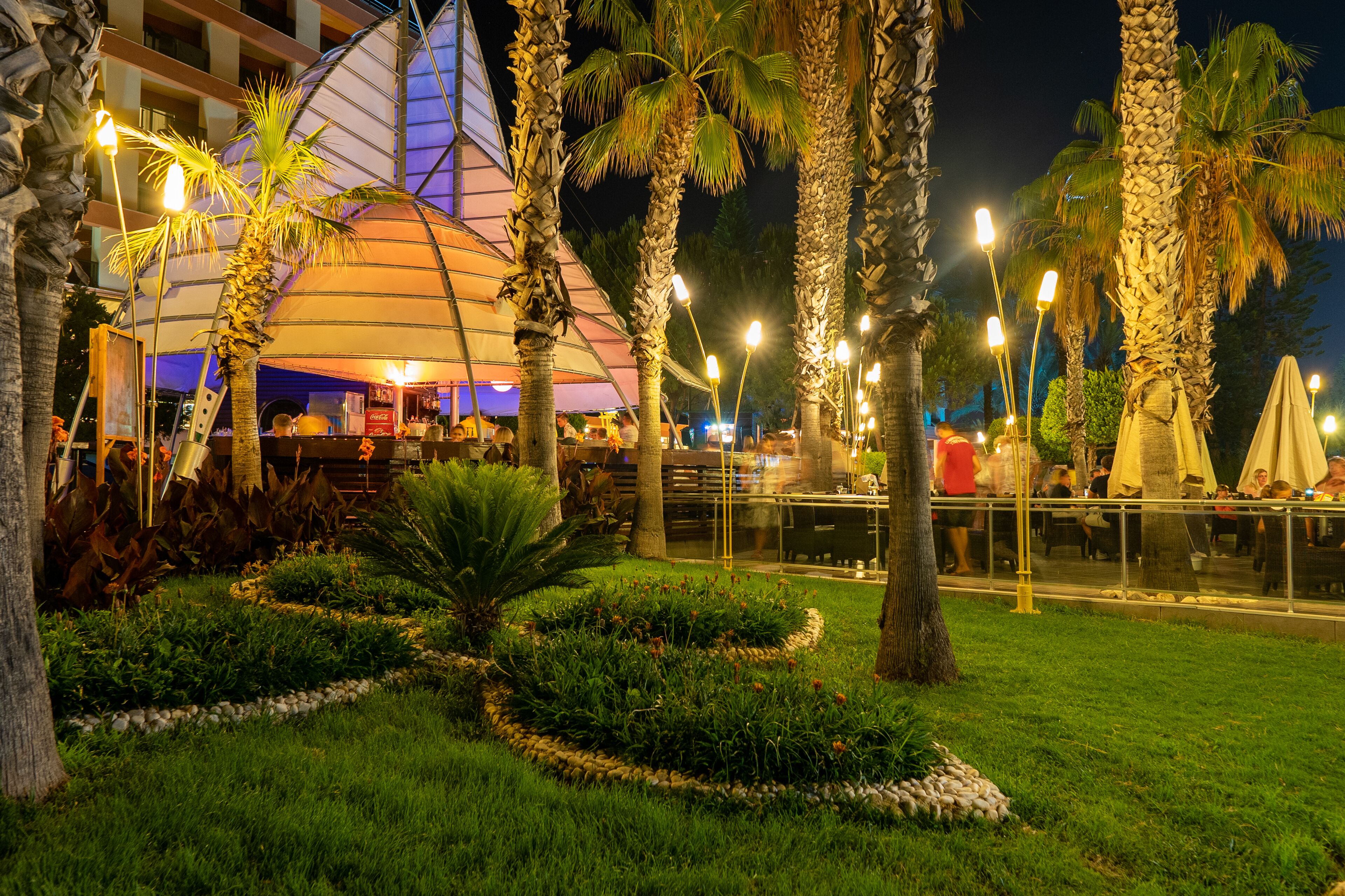 A well-lit outdoor area with palm trees, a dining area, and a building with a distinctive roof structure.