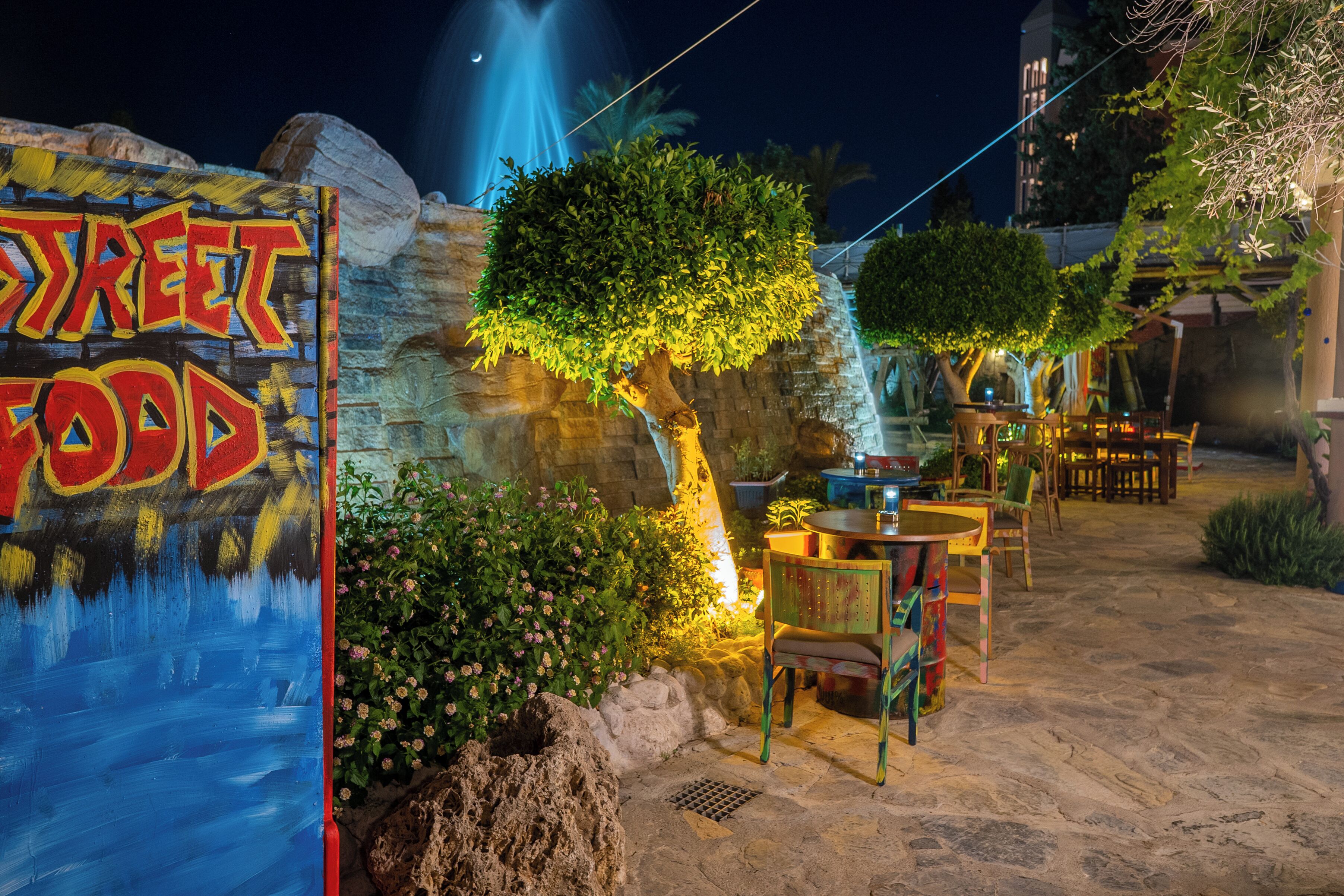 Un espace de restauration en plein air avec un panneau indiquant « Street Food », des tables, des chaises et une cascade décorative.