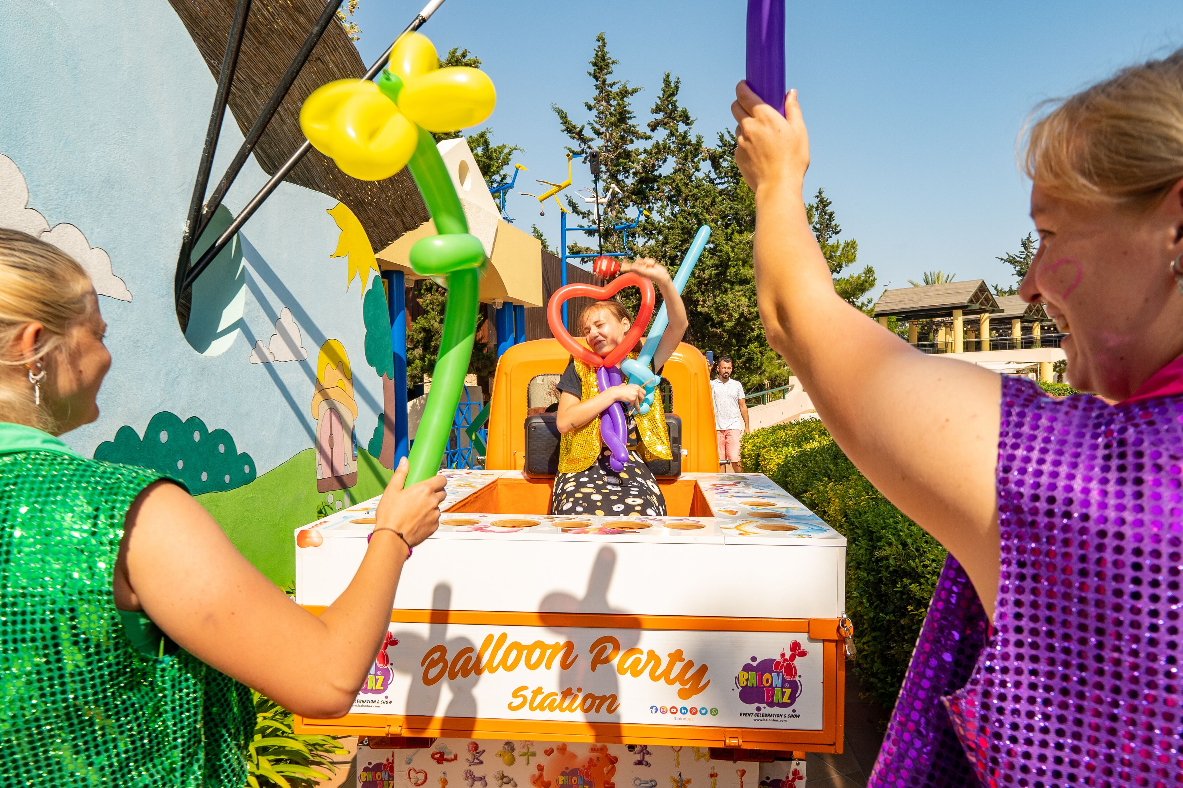 Een ballonnenkraam op een kermis, met een kleurrijke presentatie en een bordje met de tekst 'Balloon Party Station'.
