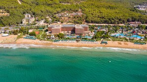 An aerial view of a resort with a large building, beach, and surrounding forested hills. - AQI Pegasos Royal - All inclusive (Alanya)