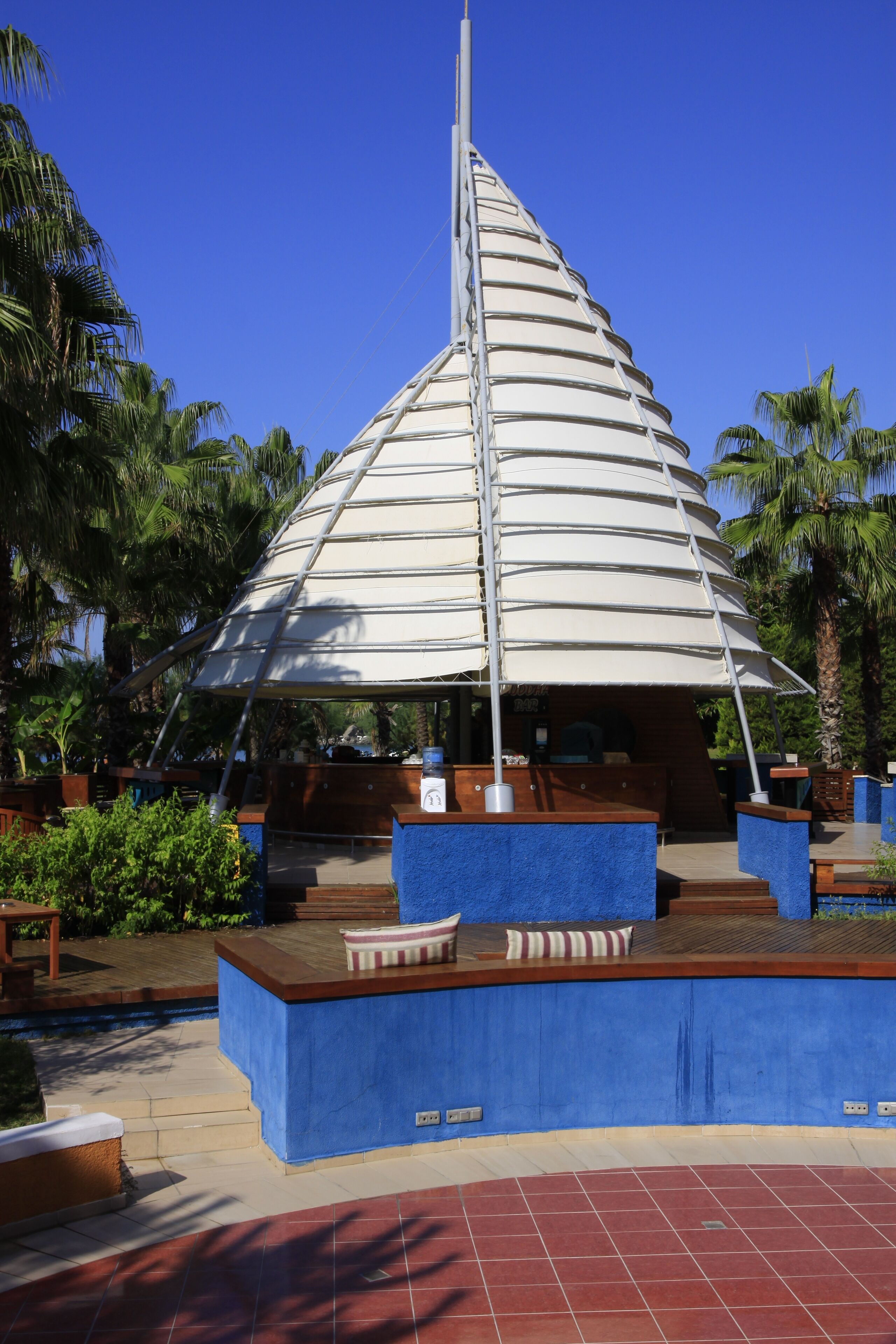 A modern, multi-level structure with a sloped roof, surrounded by palm trees and a blue-tiled floor.