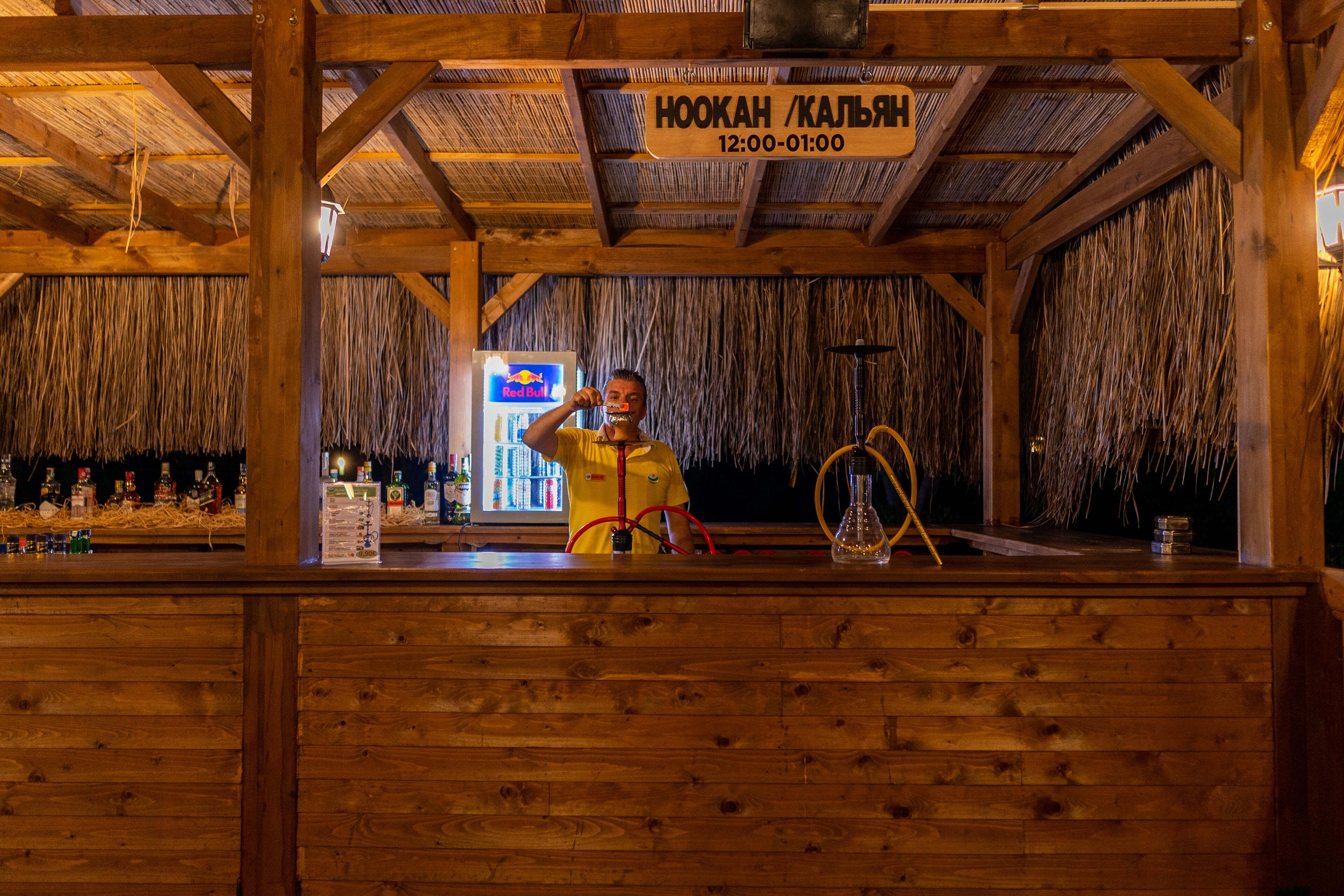 Zona de bar con barra de madera, techo de paja, letrero de Red Bull e indicaciones del horario del salón de hookah.