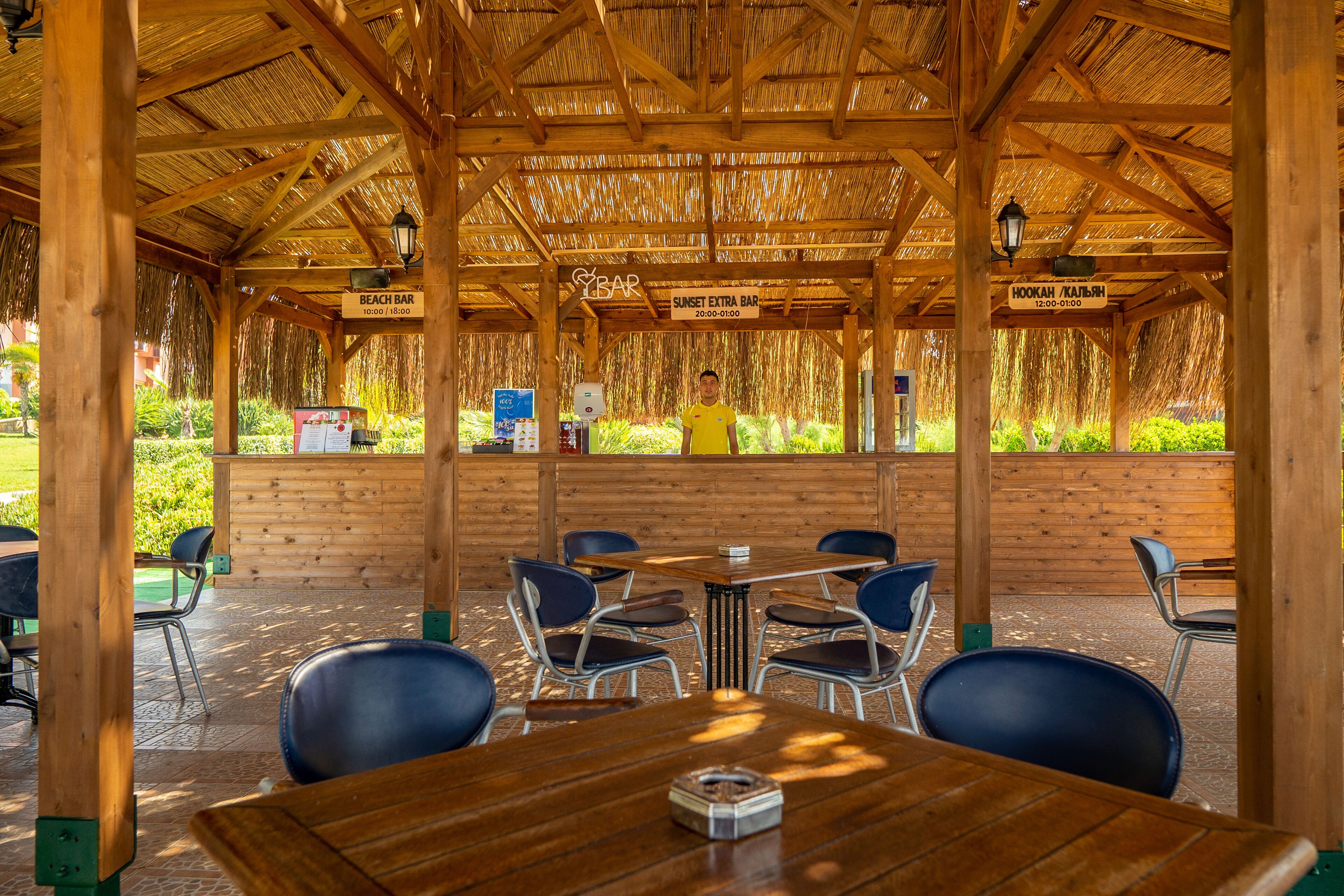 Un espacio de estar al aire libre techado con mesas y sillas de madera, un letrero de bar de playa y una persona al fondo.