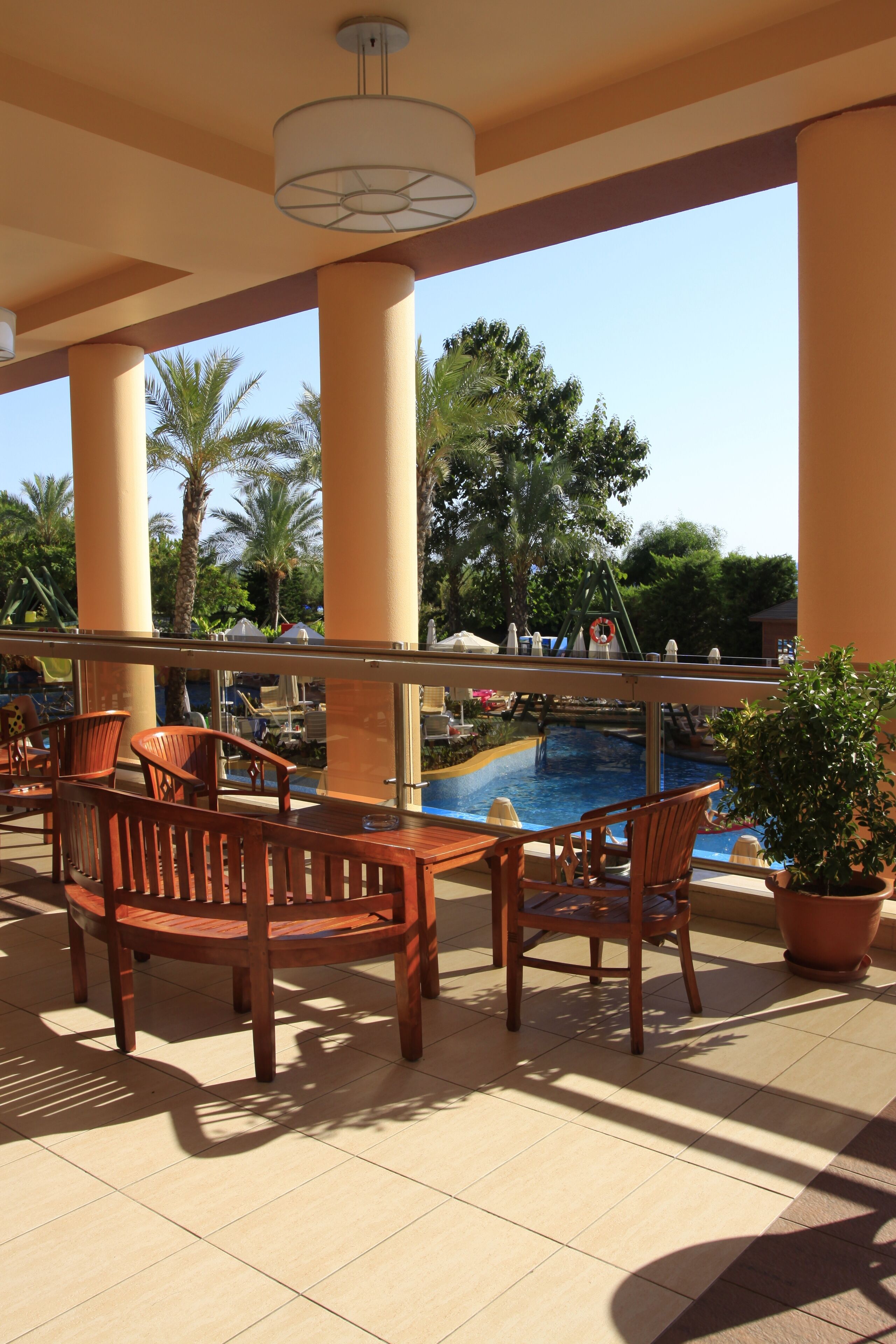 Zona junto a la piscina con sillas y una mesa de madera, rodeada de palmeras y un cielo despejado.