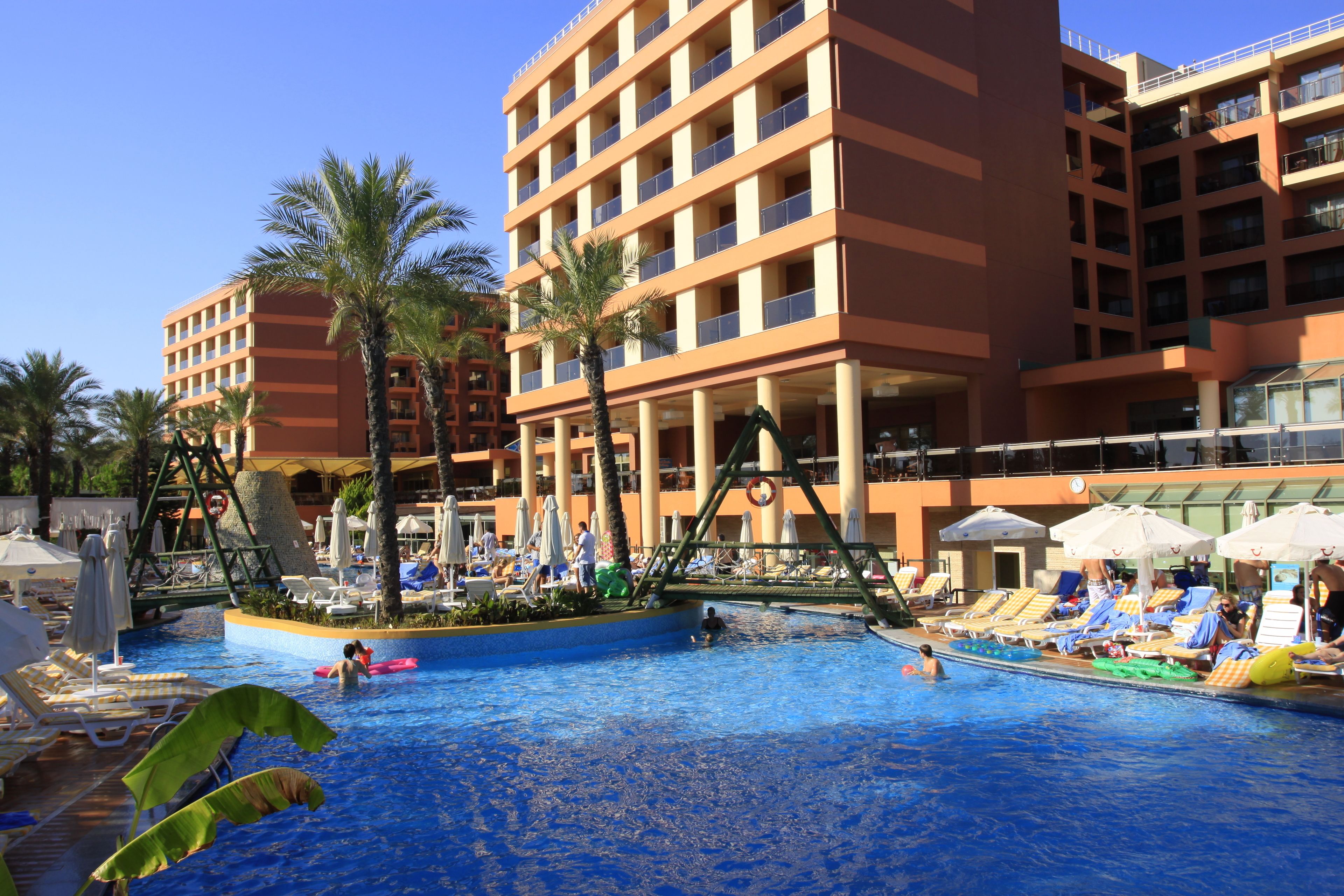 A hotel pool area with lounge chairs, umbrellas, and palm trees.
