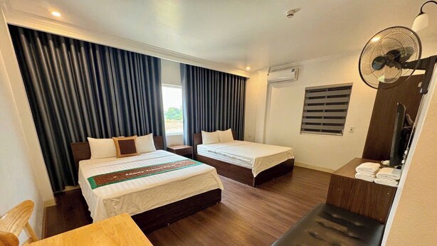 A hotel room with two beds, a wooden table, a bench, a fan, and a window with curtains. - Family Transit Hotel (Hanoi)