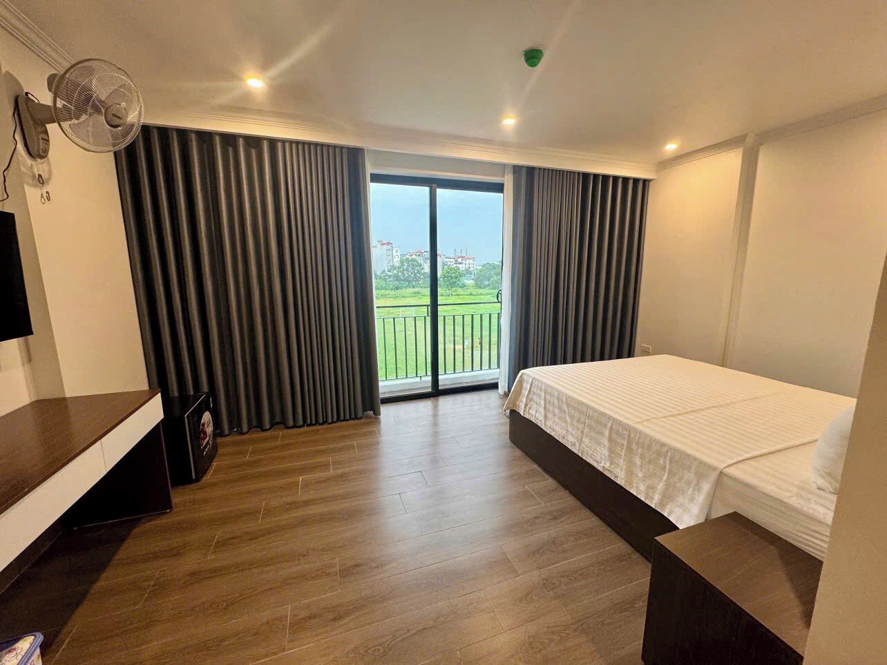 Una habitación de hotel con cama, televisión, ventilador y balcón con cortinas.