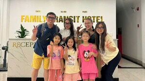 A family group posing in front of a reception desk at a hotel with a sign indicating it is a family transit hotel. - Family Transit Hotel (Hanoi)