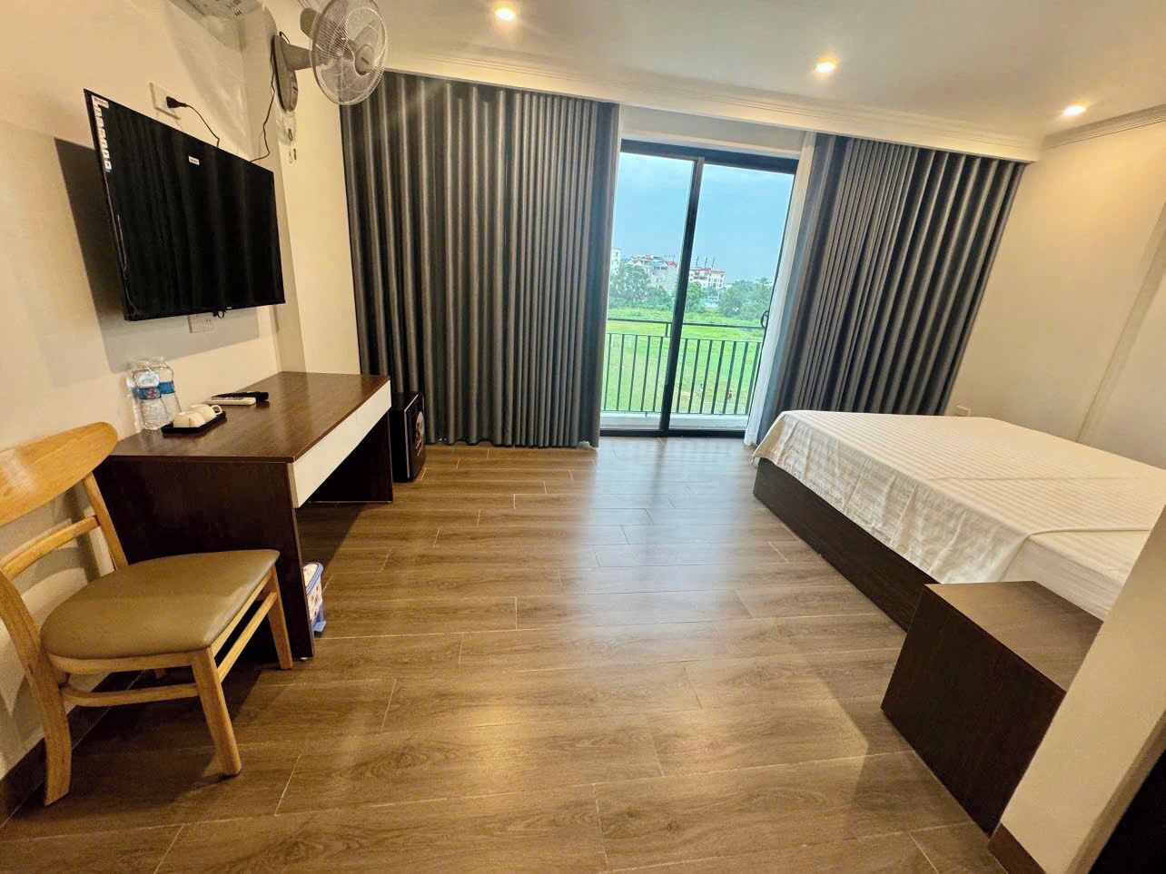 Habitación de hotel con cama, escritorio, silla, televisor y balcón con cortinas.