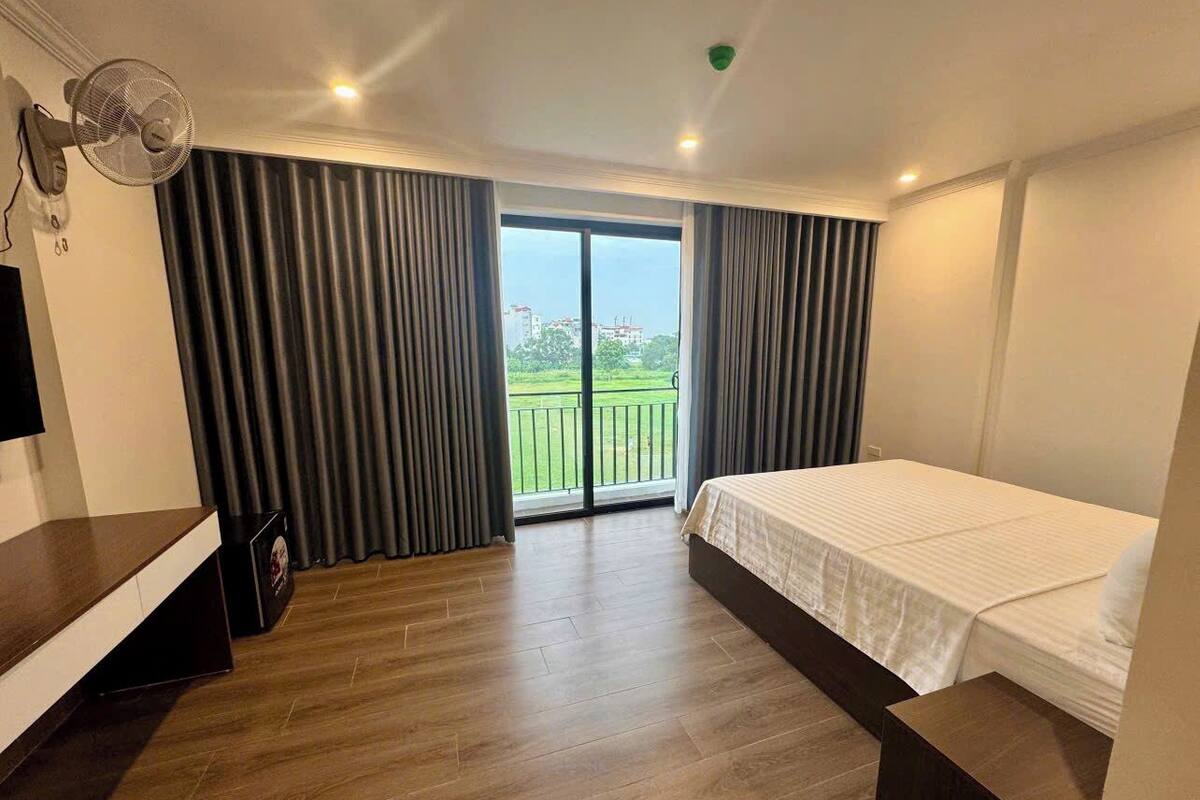 A hotel room with a bed, a TV, a fan, and a balcony with curtains.