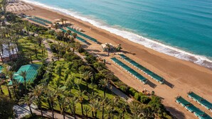 Vista aérea de un complejo turístico en la playa, con hileras de sillas de playa, una torre de salvavidas y una zona de piscina.