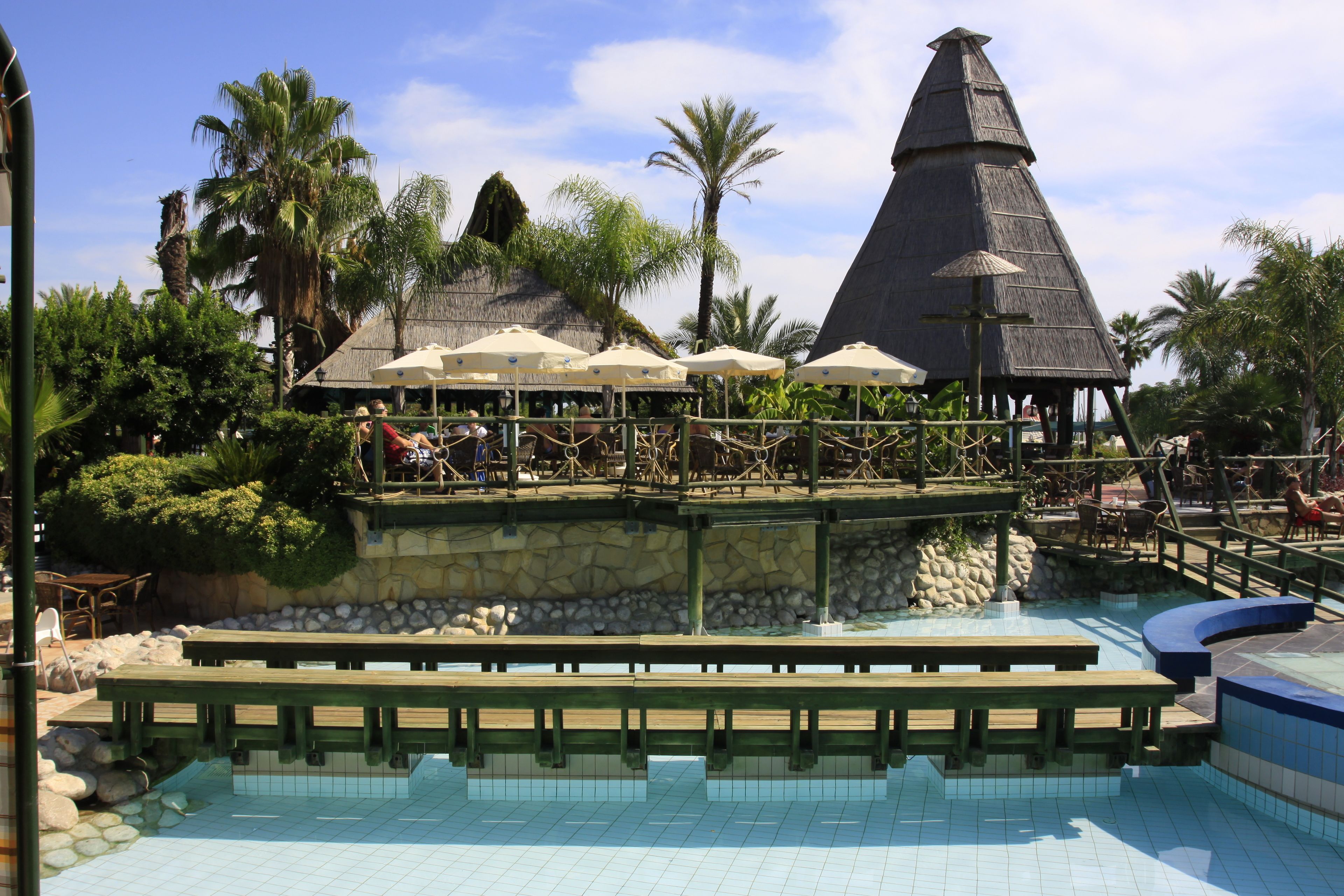 A pool area with wooden benches, umbrellas, and a thatched-roof structure.