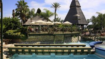 A pool area with wooden benches, umbrellas, and a thatched-roof structure.