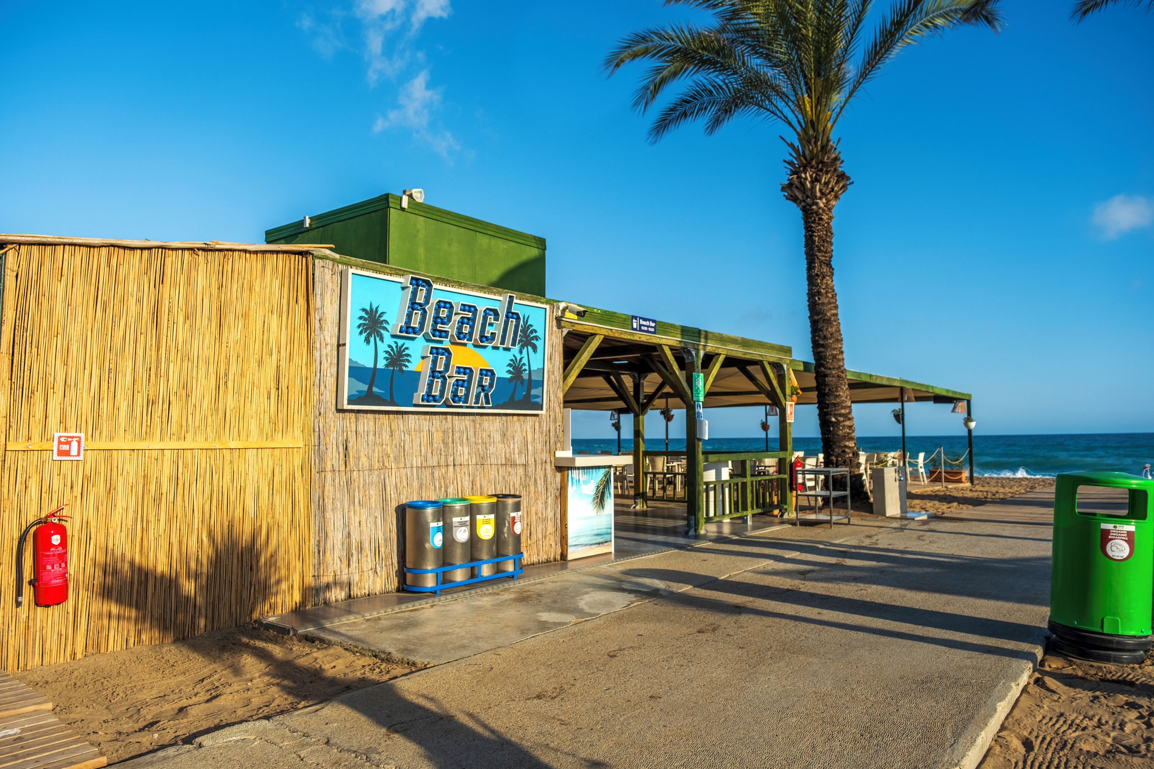 Un bar de playa con un letrero, un bote de basura y una palmera.