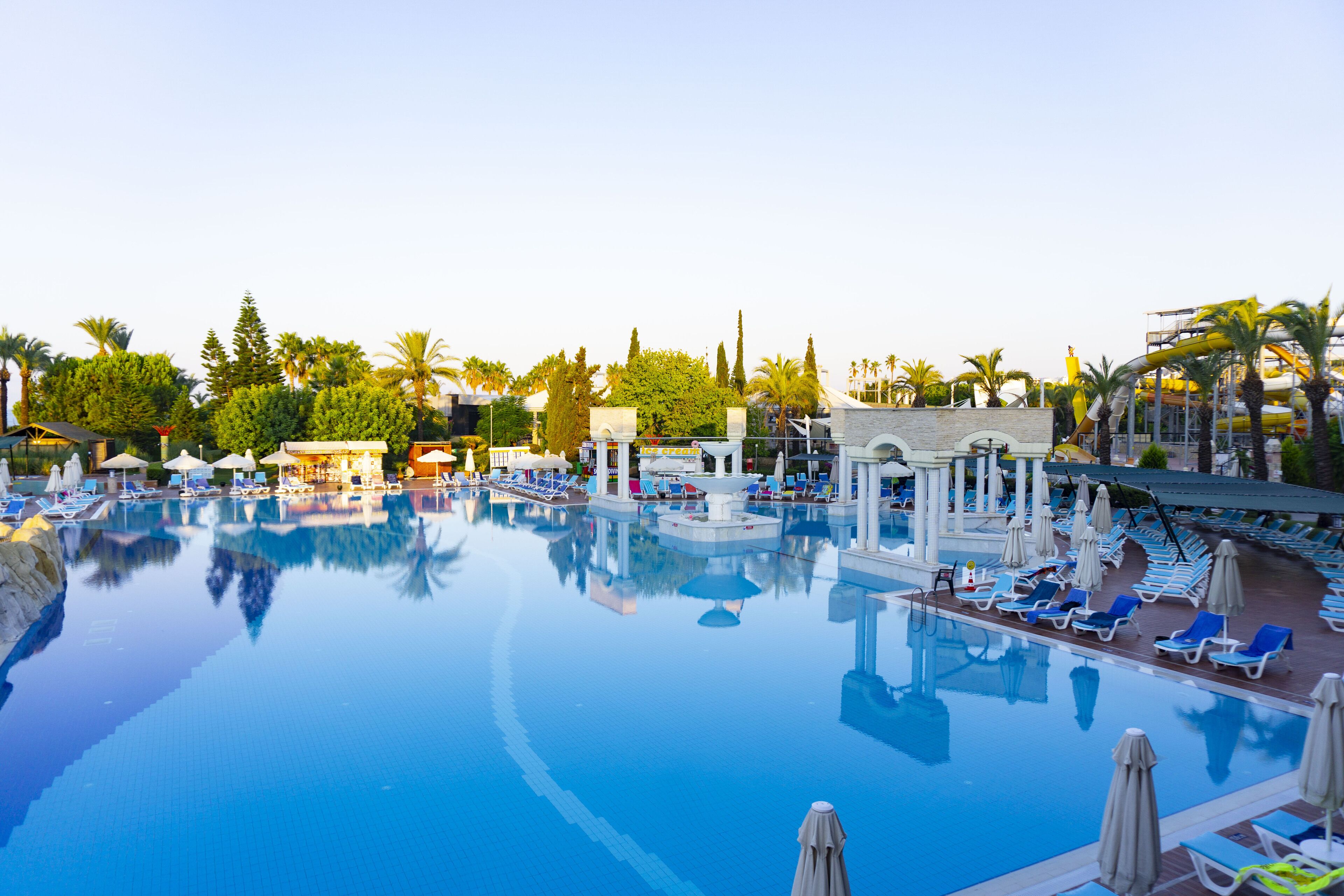 Una gran piscina con sillas de descanso y sombrillas, rodeada de palmeras y un edificio con fachada blanca.