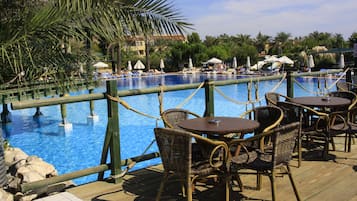 Un espace aménagé au bord de la piscine, avec des fauteuils et des tables en osier, une terrasse en bois et une vue sur la piscine et les palmiers environnants.