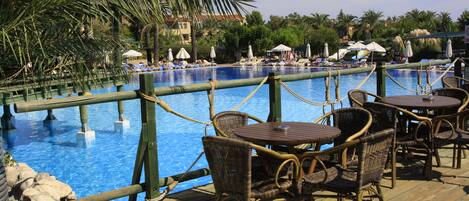 A poolside area with wicker chairs and tables, a wooden deck, and a view of the pool and surrounding palm trees.