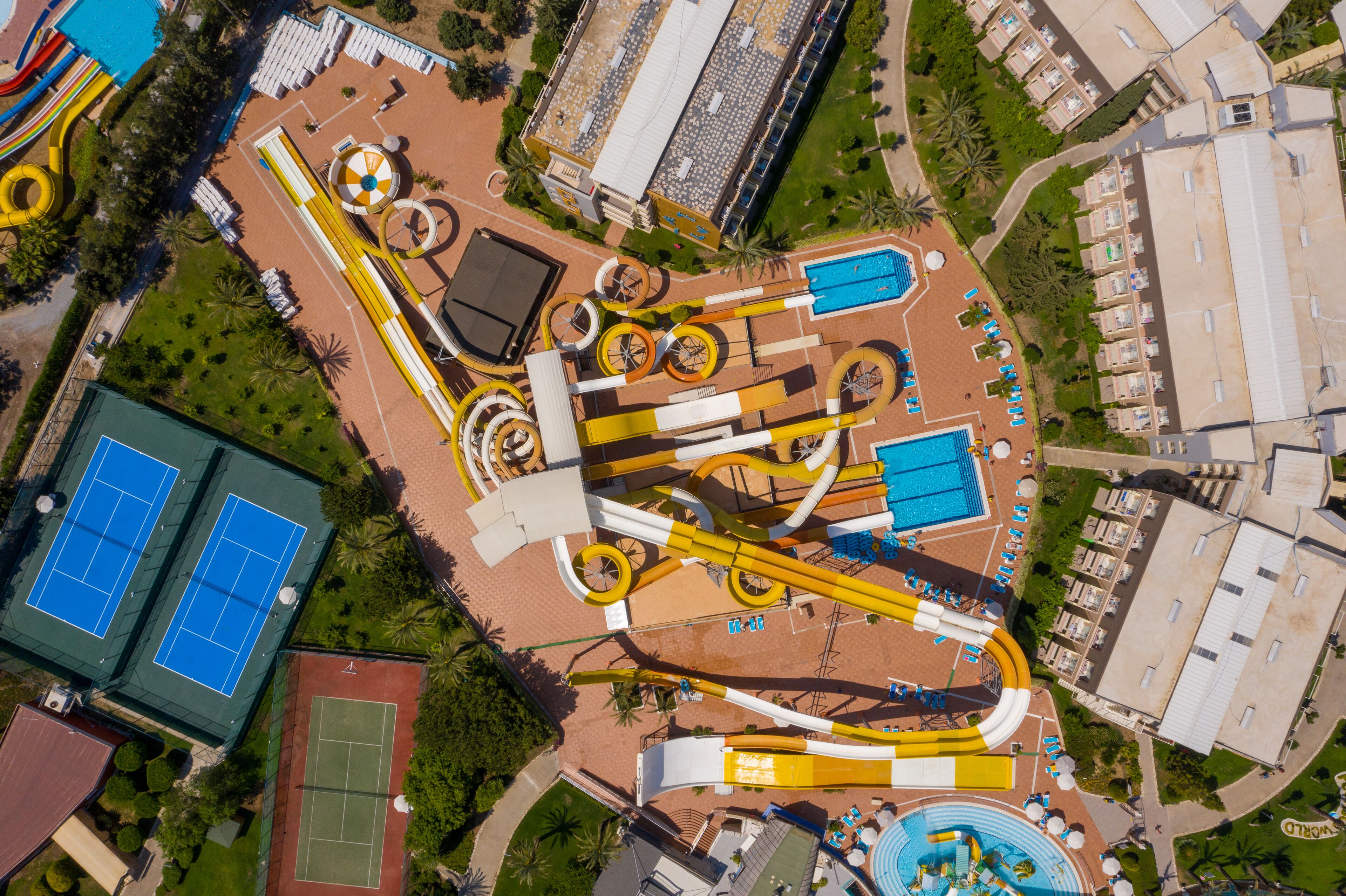 An aerial view of a resort with multiple swimming pools, water slides, tennis courts, and a large building with a flat roof.