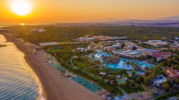 Vista aérea de un complejo turístico con una piscina grande, playa y montañas al fondo.