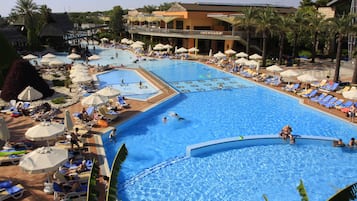 A large outdoor swimming pool with multiple lanes, surrounded by sun loungers and umbrellas, and adjacent to a building with balconies.