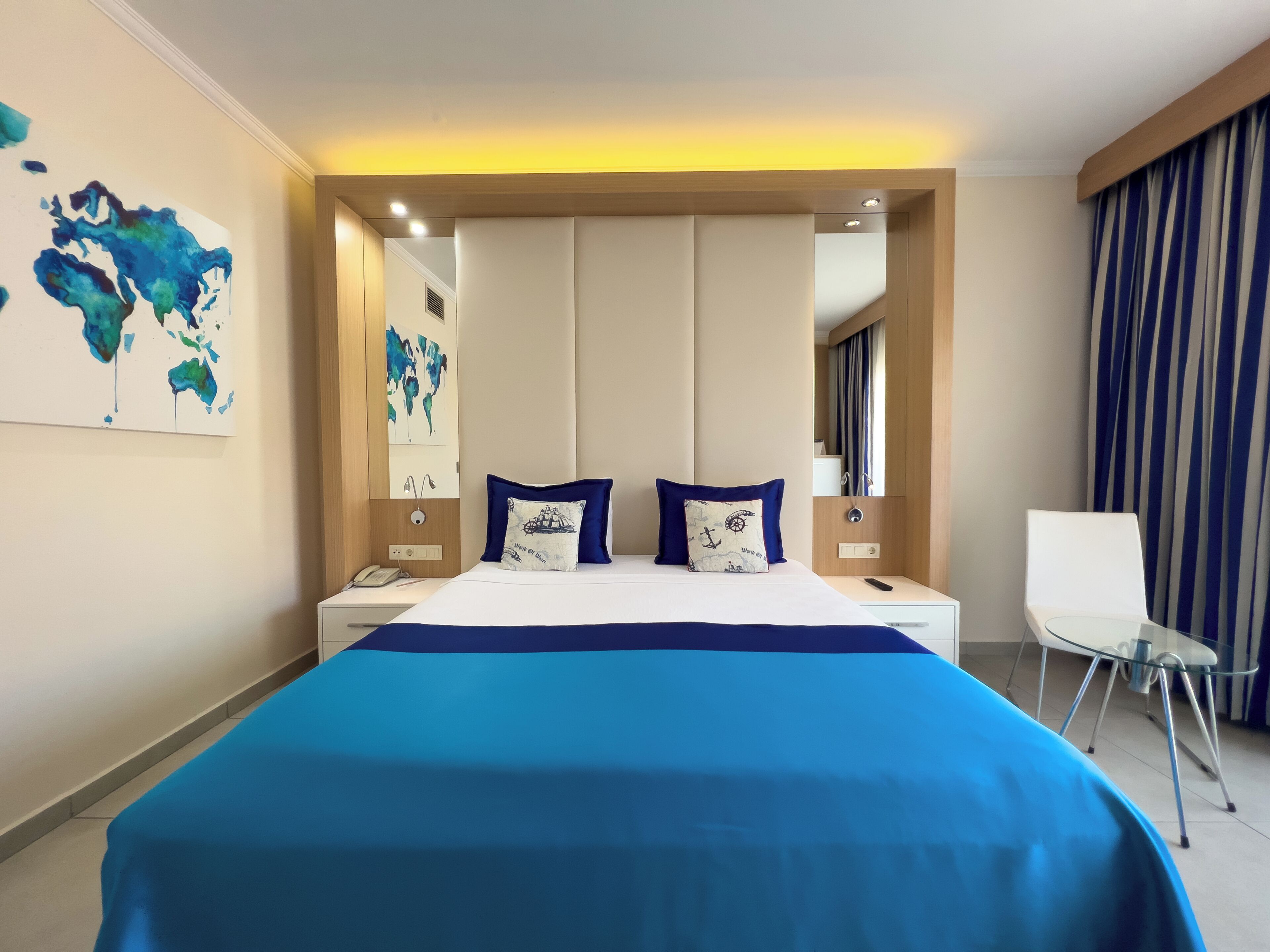 A modern hotel room with a large bed, two pillows, a bedside table, a chair, and a world map artwork on the wall.