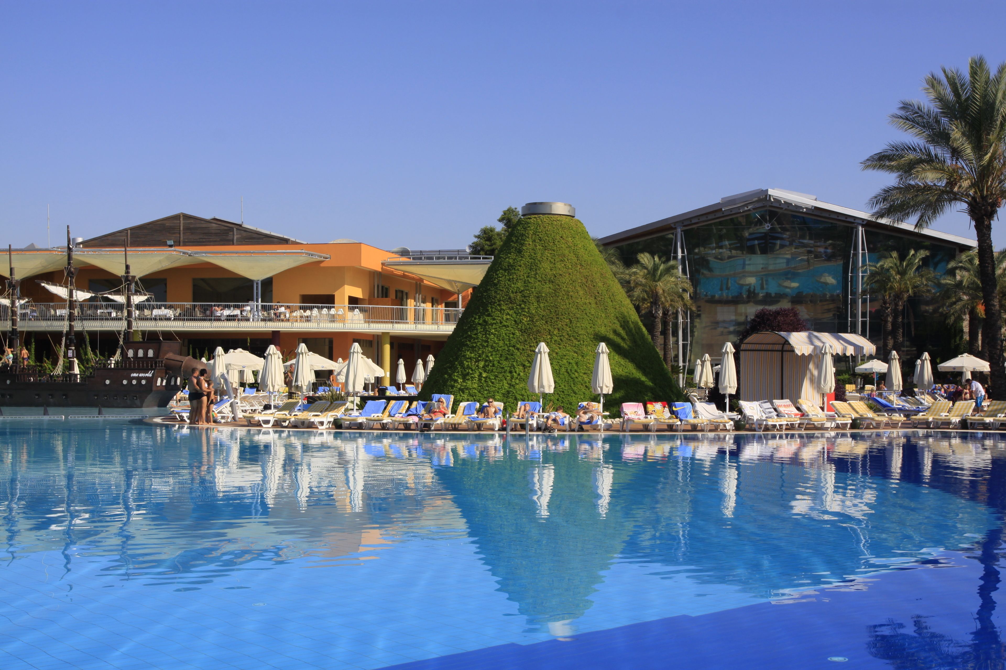 A swimming pool with lounge chairs and umbrellas, a green conical structure, and a building with a glass facade.