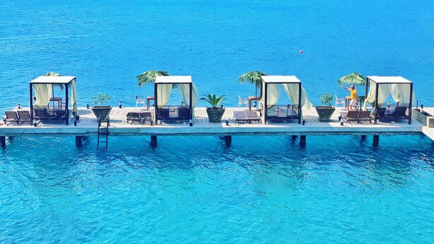 Une plateforme flottante dotée de quatre espaces de repos abrités, chacun comprenant une chaise et une petite table, et offrant une vue sur une étendue d’eau d’un bleu limpide.