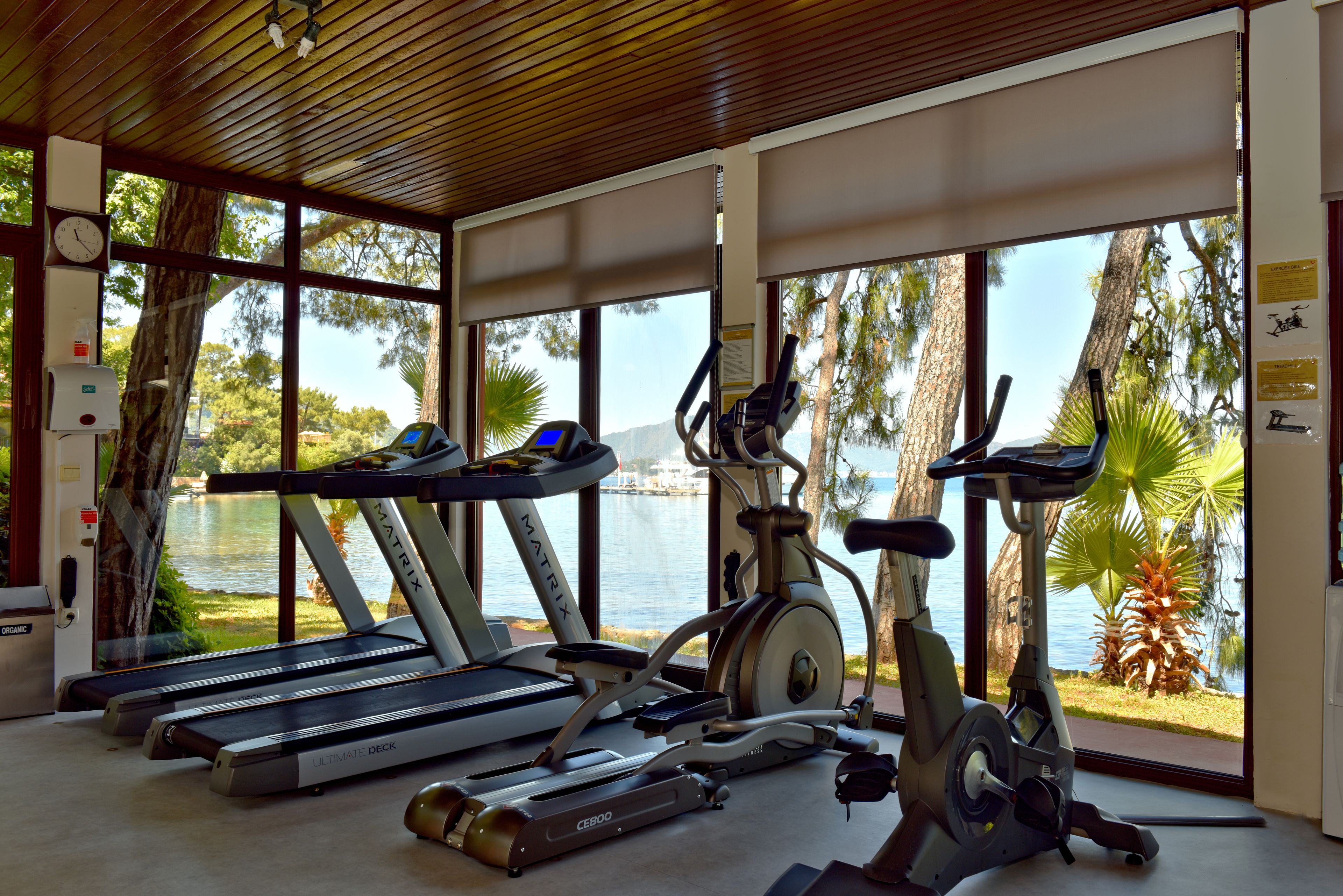 A gym with treadmills, an elliptical, and a stationary bike, overlooking a lake with trees and a building in the background.