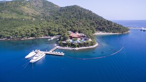 Une vue aérienne d’une zone côtière avec une petite île, sur laquelle se trouve un quai avec des bateaux, le tout étant entouré d’eaux d’un bleu limpide et de collines verdoyantes.