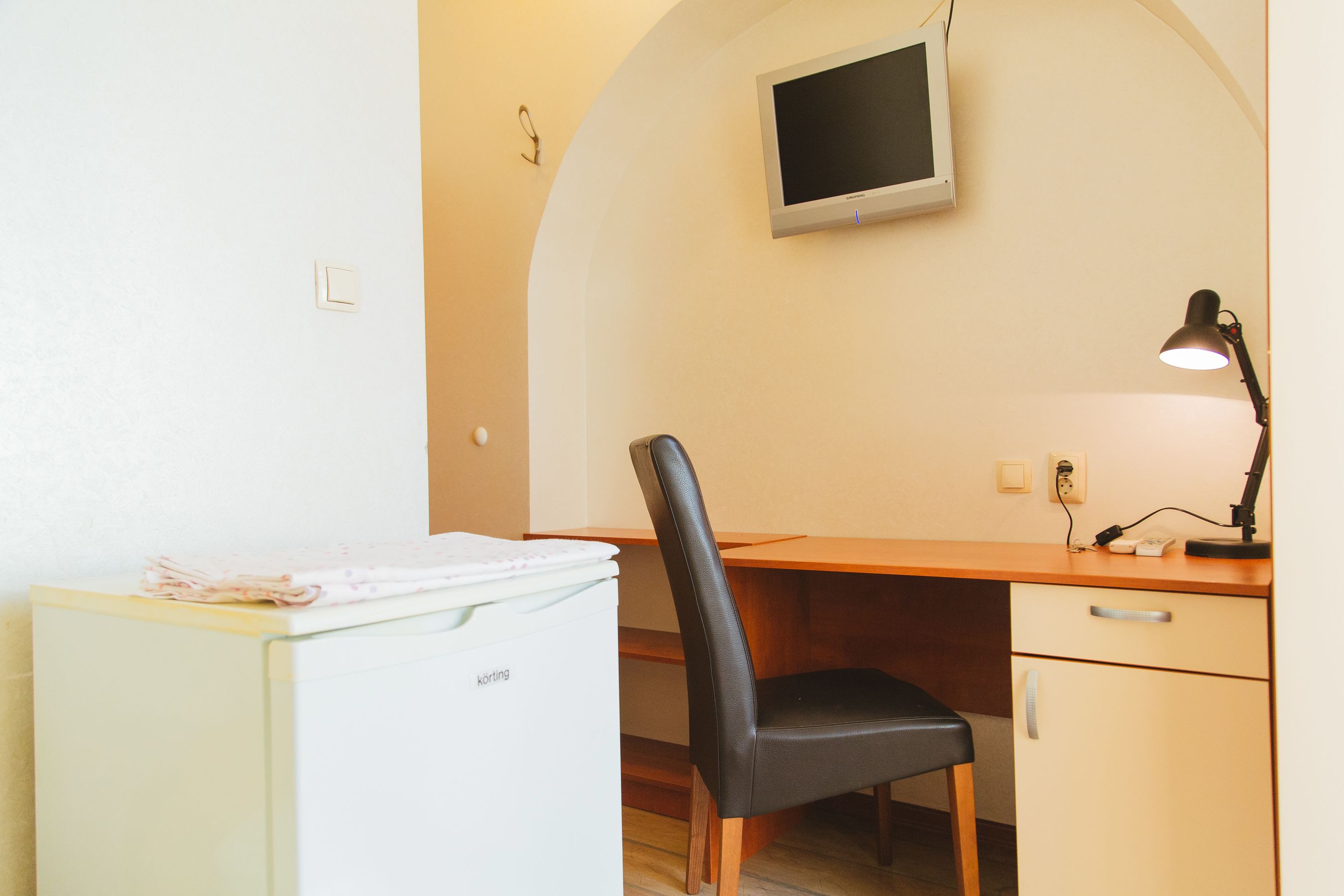 Une pièce de taille modeste, comprenant un bureau, une chaise et un petit réfrigérateur blanc.