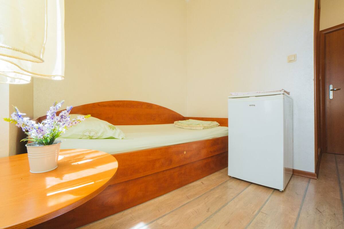 Una habitación compacta con una cama, una mesita con una planta y un refrigerador.