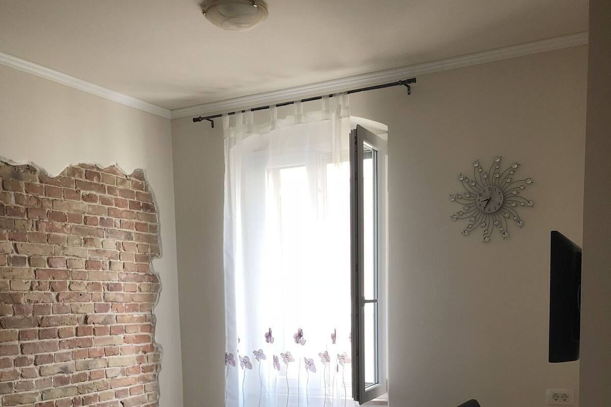 Una habitación con pared de ladrillo, sofá verde, escritorio de madera y cortina blanca con motivos florales.