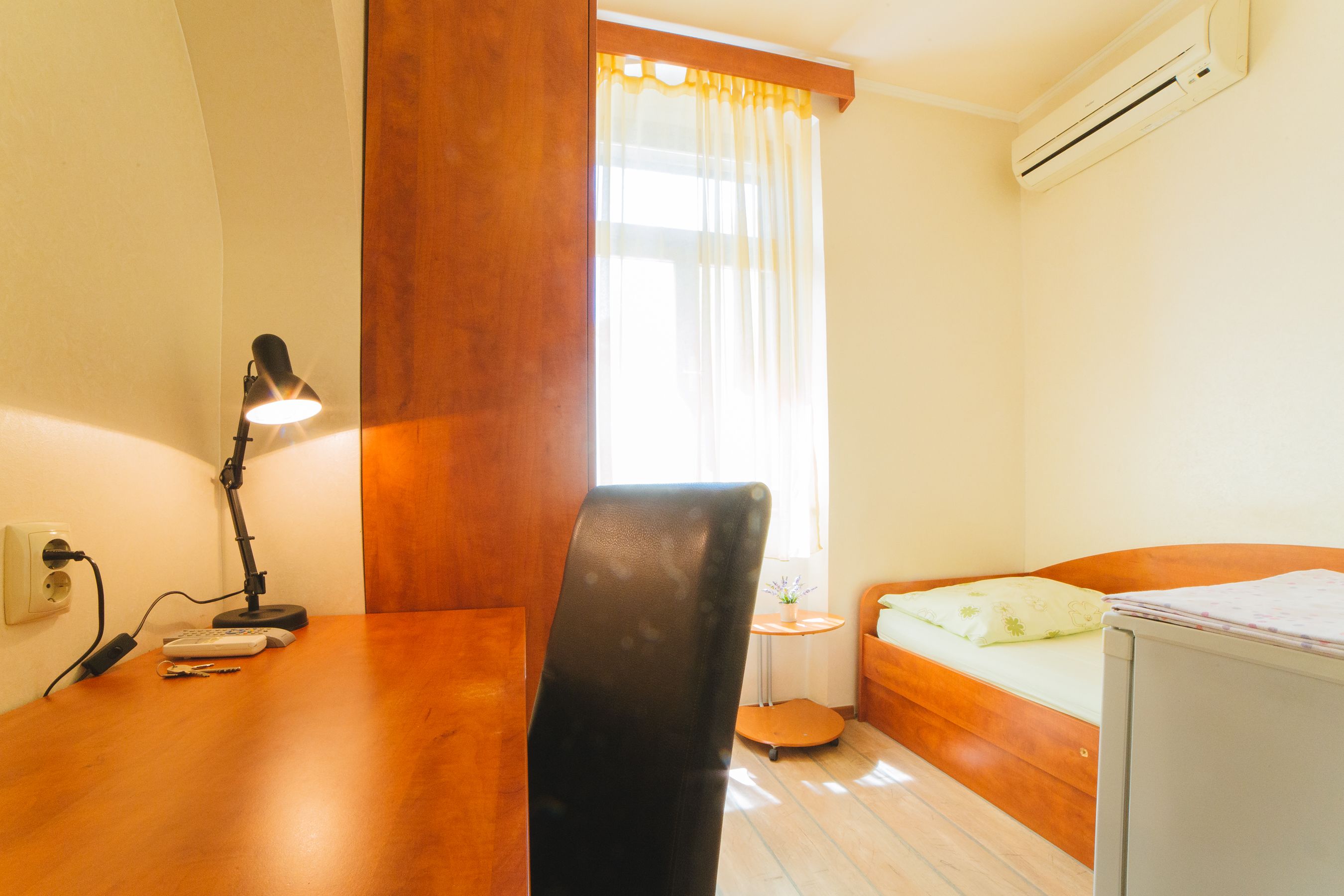 Una habitación pequeña y luminosa con un escritorio de madera, una silla, una cama y una ventana con cortinas.