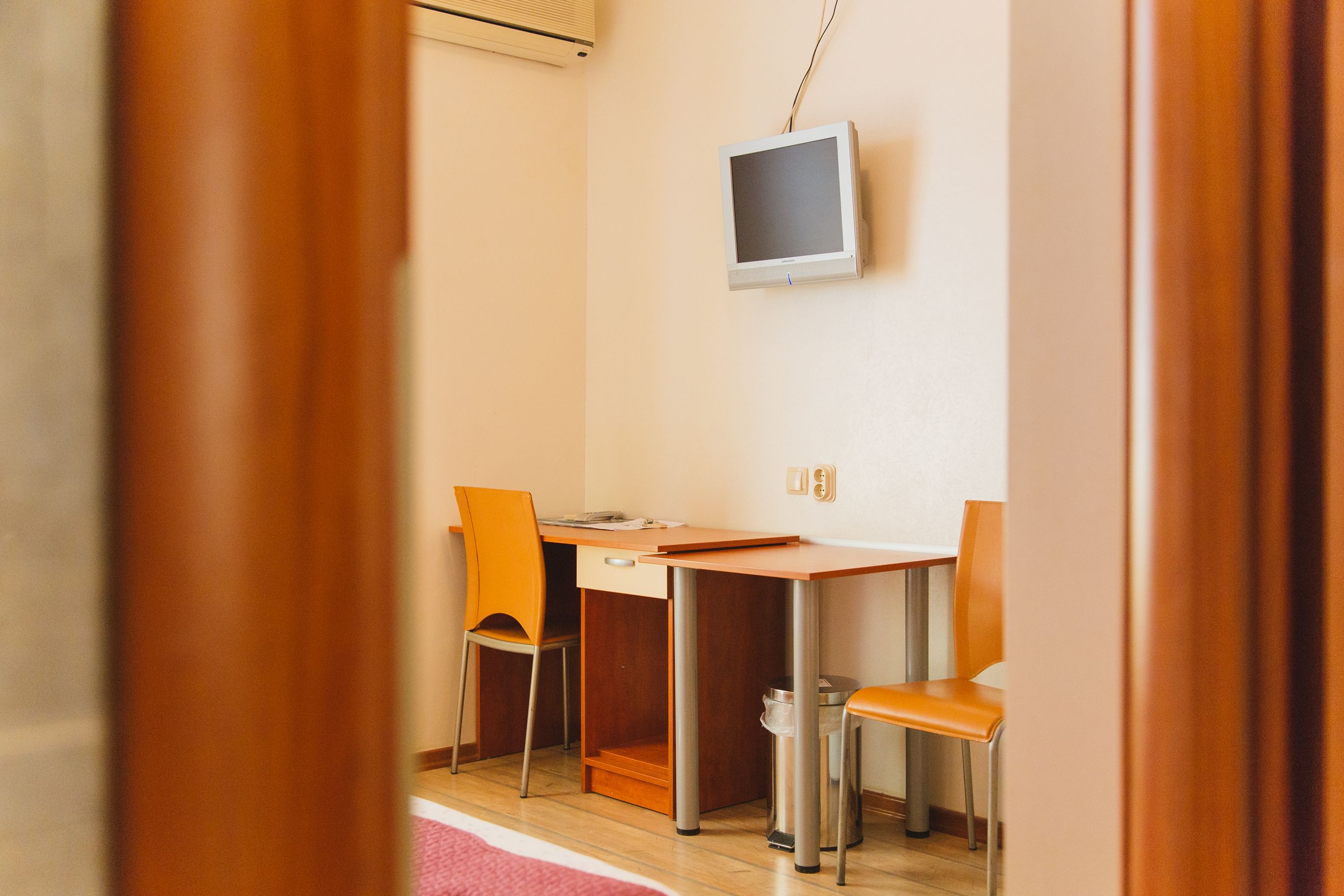 A room with a wooden desk, two chairs, a television on the wall, and a trash can.