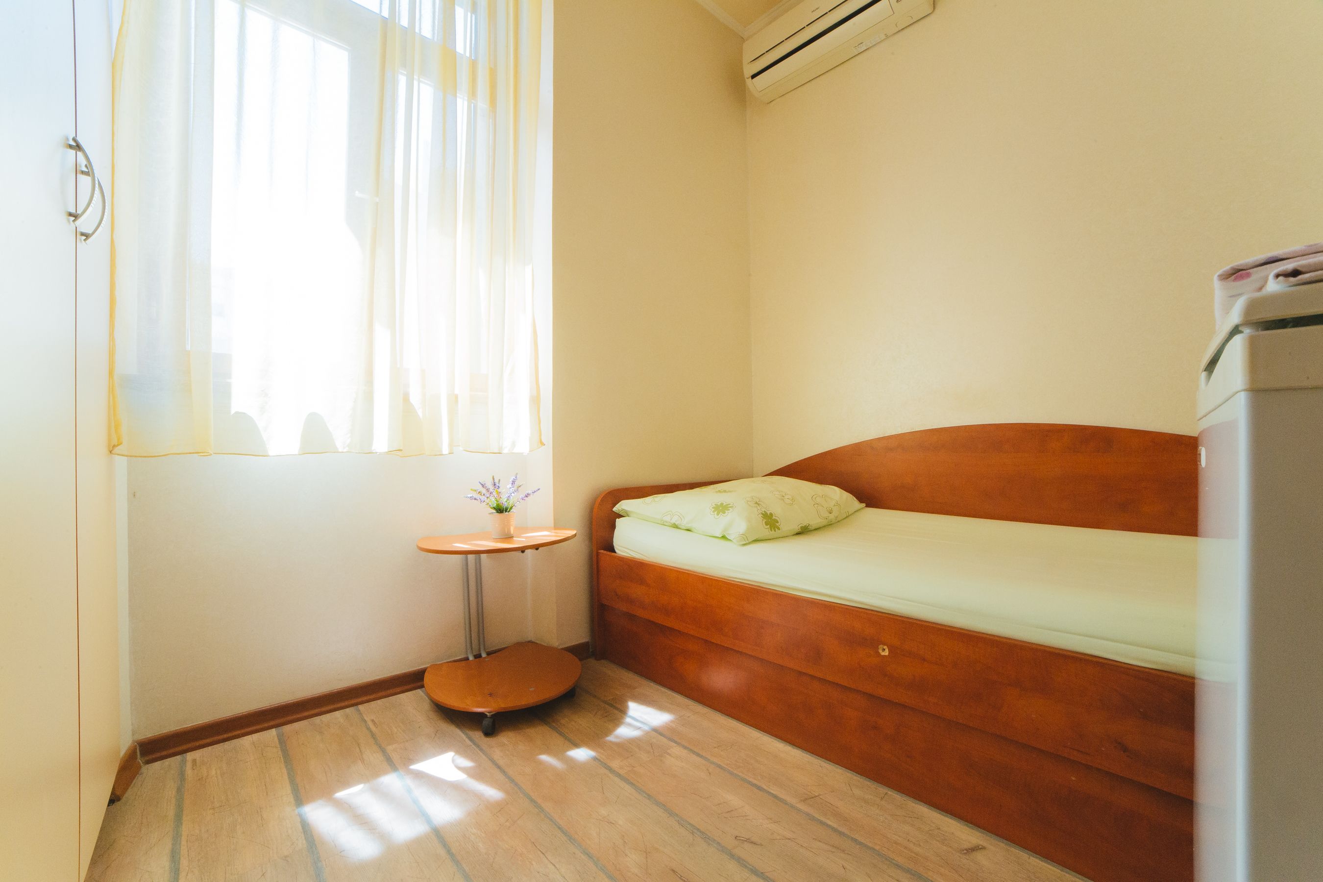 Ein kleiner, kompakter Raum mit einem hölzernen Bett, einem kleinen runden Tisch und einem Fenster mit durchsichtigen Vorhängen.