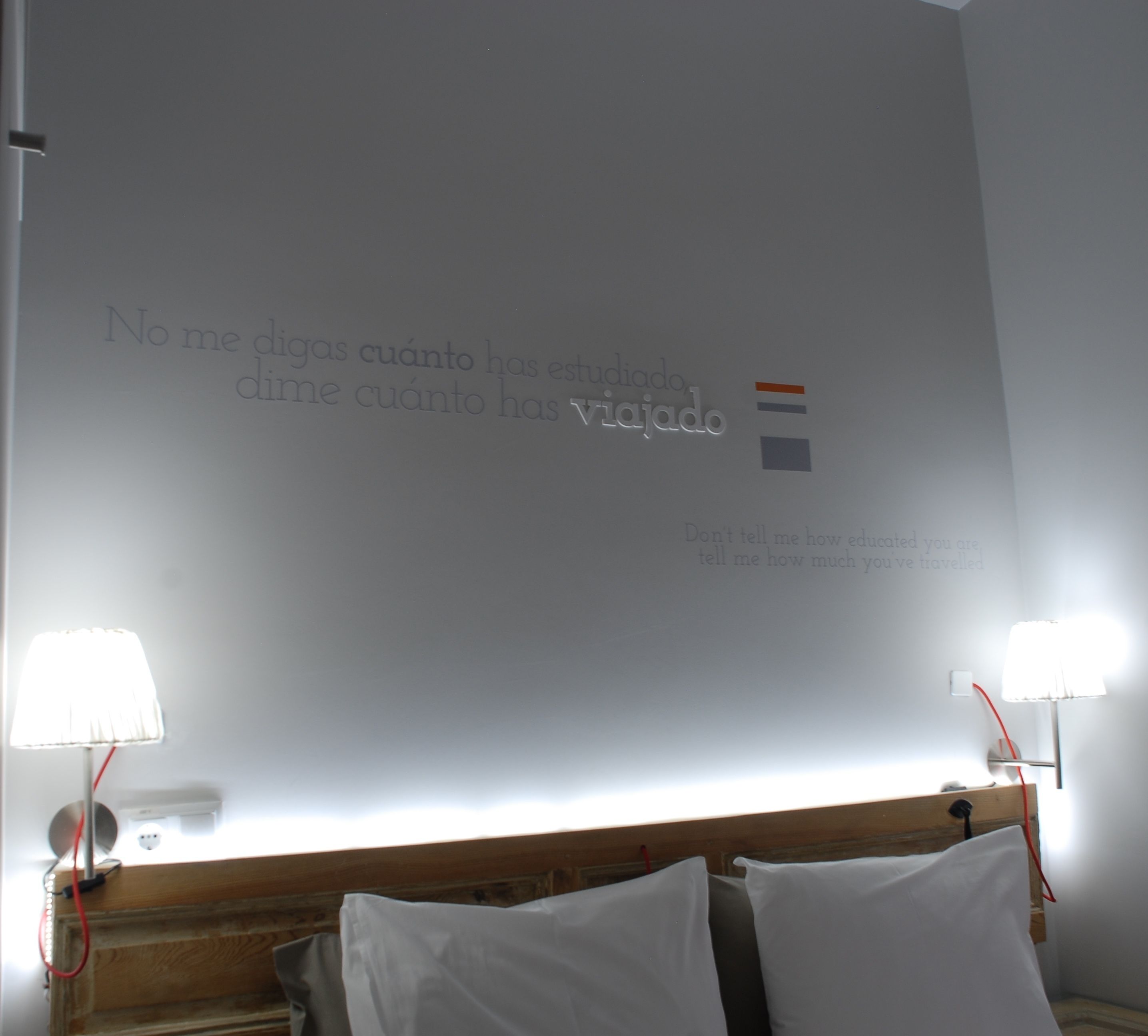 Habitación de hotel con una cama, dos lámparas de noche y una pared con una cita motivacional en español.