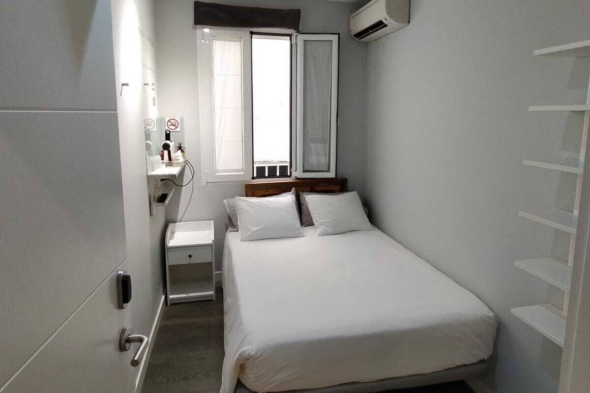 Camera d'albergo compatta con un letto singolo, un comodino, una piccola cassettiera, una finestra con tende e un condizionatore d'aria.