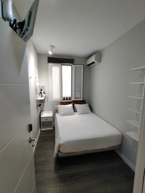 A compact hotel room with a single bed, a bedside table, a small dresser, a window with curtains, and an air conditioning unit. - Diezmadrid Hostal (Madrid)