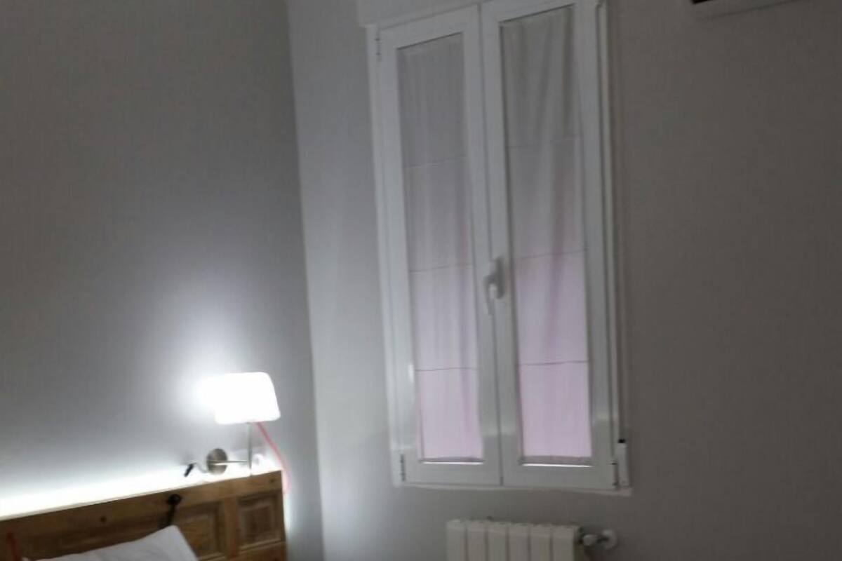 Una camera d'albergo con un letto, un comodino con lampada, una finestra con tende e un condizionatore d'aria.