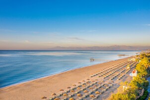 Een strand met rijen ligbedden en parasols, een pier die het water in reikt, en een vlag aan de rechterkant.