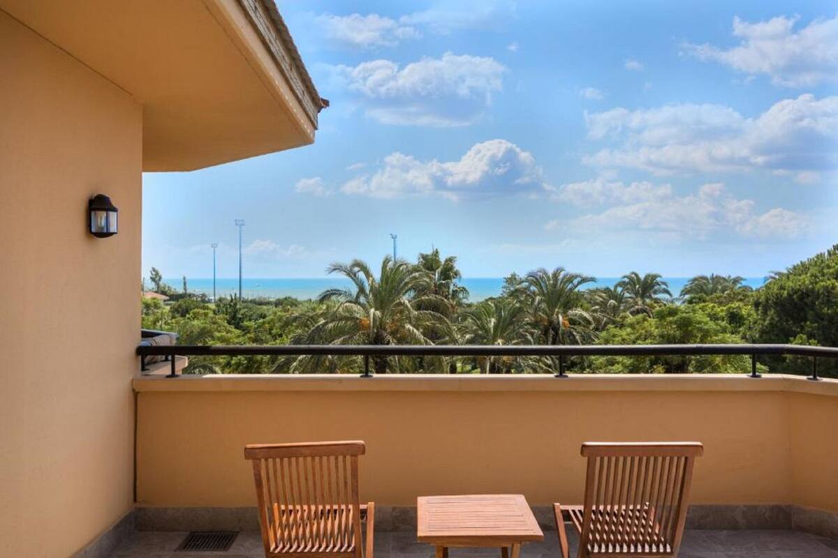 A balcony with two wooden chairs and a small table, overlooking a lush green landscape and the sea.